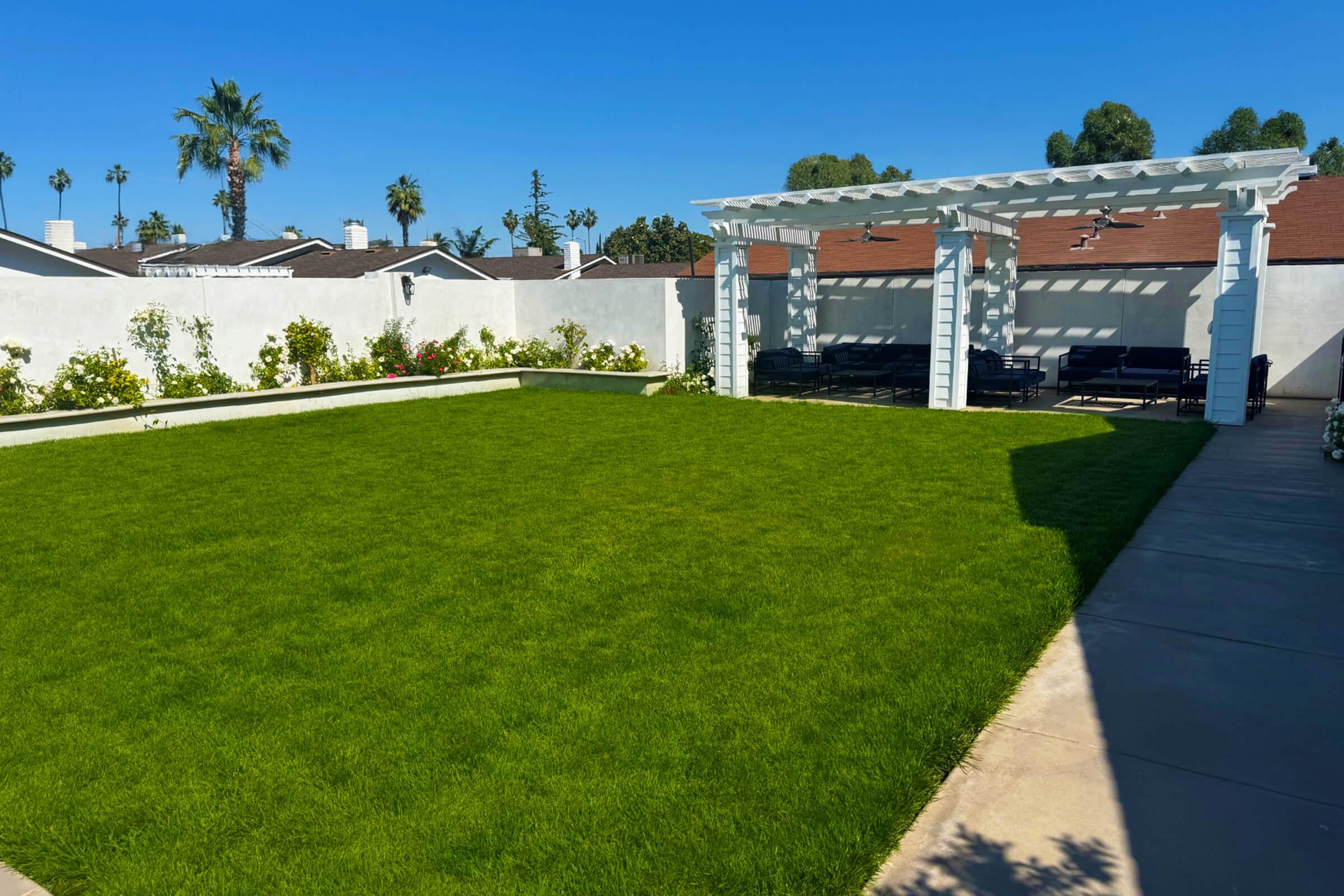 A spacious backyard featuring lush green grass, bordered by white walls and colorful flowers. In the background, a white pergola provides shade over a seating area with dark outdoor furniture, all under a clear blue sky with palm trees in the distance.