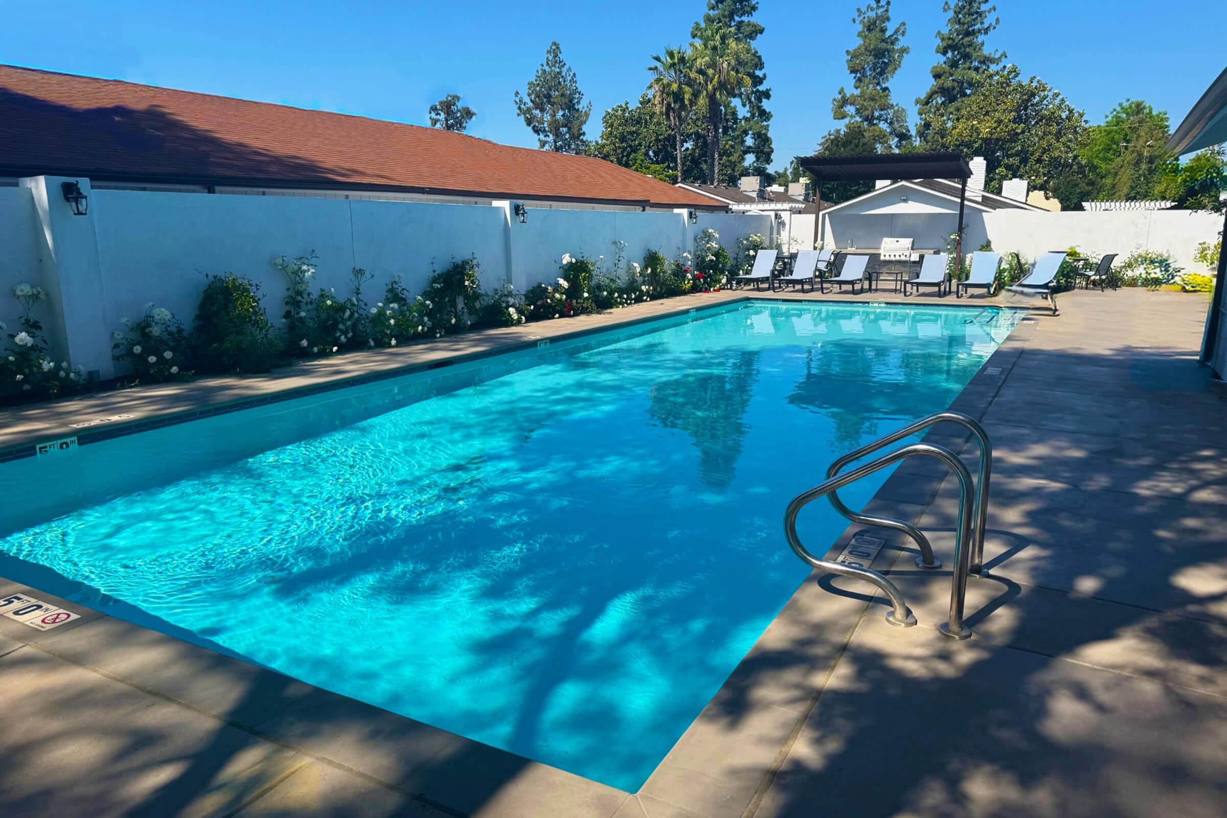 A clear blue swimming pool surrounded by a patio with lounge chairs. Numerous trees and shrubs line the area, adding greenery. The pool area is enclosed by a white wall, and there is a shaded seating area at one end, providing a relaxing outdoor space.