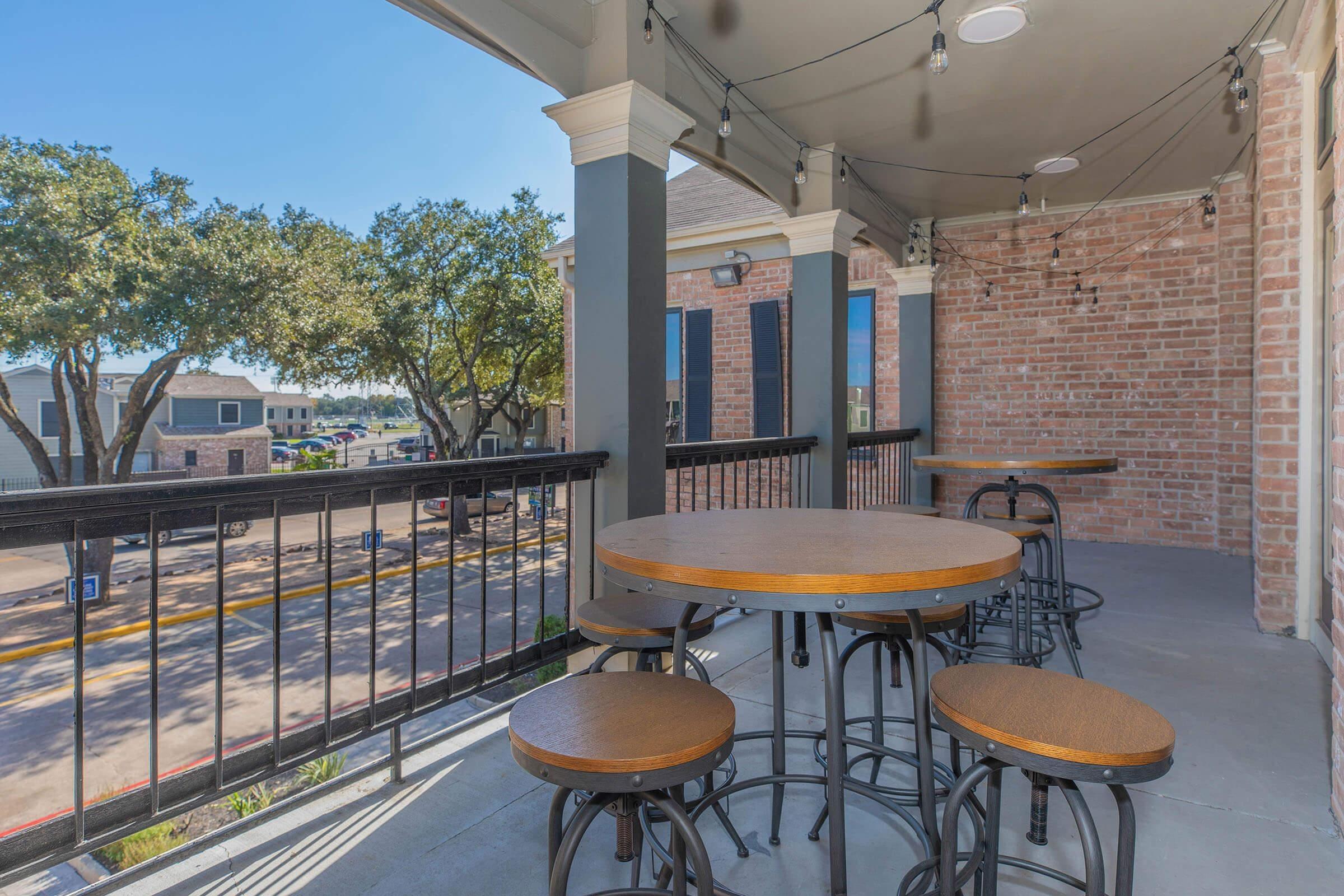 A spacious balcony with a wooden round table and four black metal chairs. The balcony features a railing and string lights, surrounded by trees. In the background, there are buildings and a clear blue sky visible, creating a relaxed outdoor atmosphere.