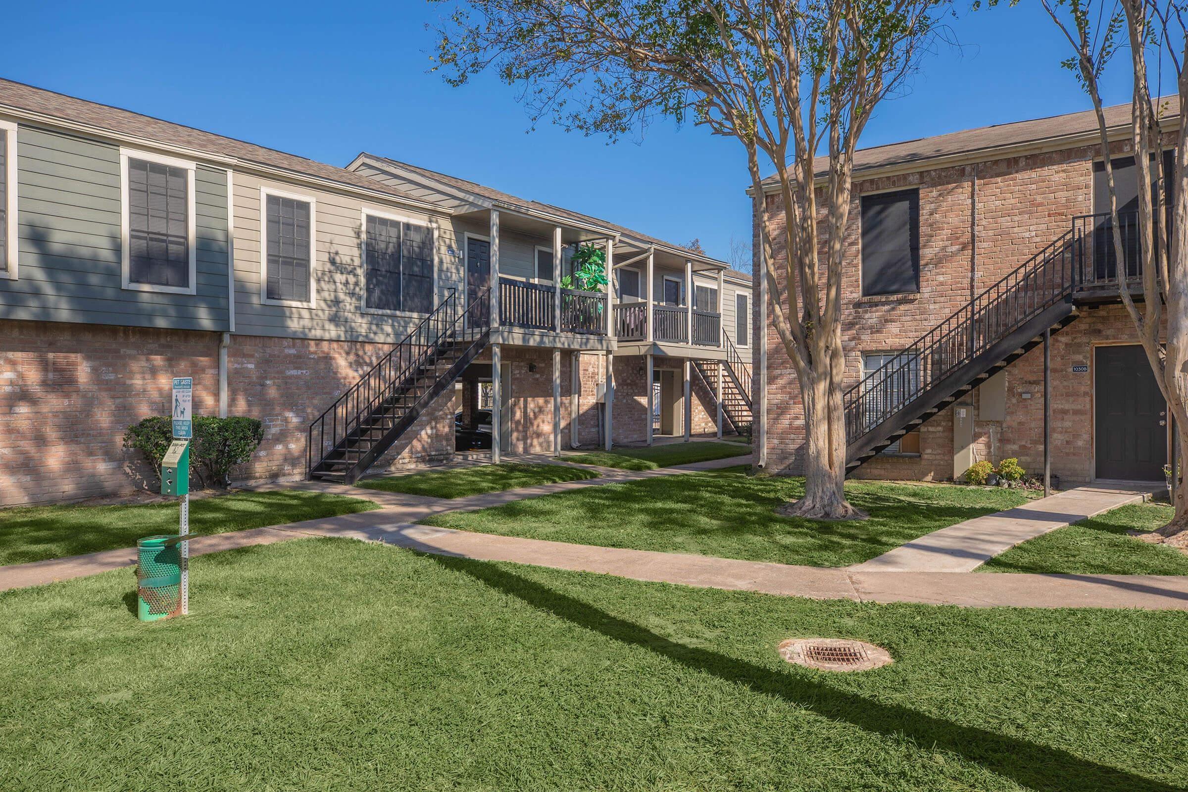 Two multi-story apartment buildings surround a grassy courtyard. The left building features a green door and multiple balconies, while the right building has a staircase leading to the upper level. Trees are visible around the area, and a sidewalk runs through the lawn, creating a welcoming outdoor space.