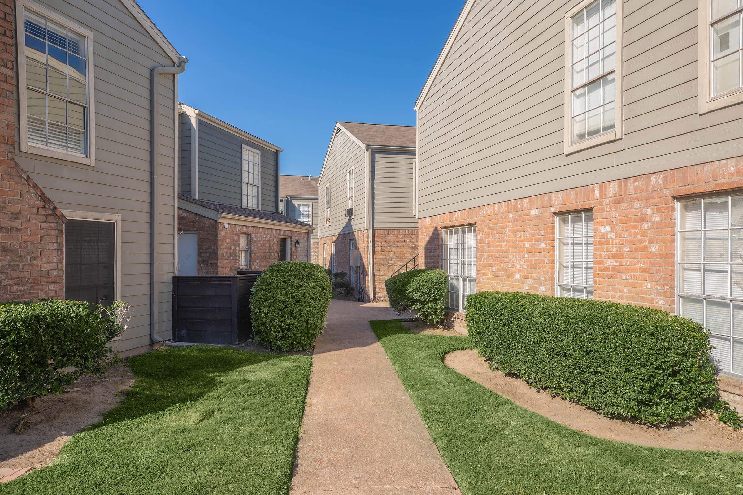 A clear pathway runs between two sets of apartment buildings, with well-maintained grass and neatly trimmed hedges on either side. The buildings have a mix of brick and siding exteriors under a bright blue sky, creating a pleasant residential atmosphere.