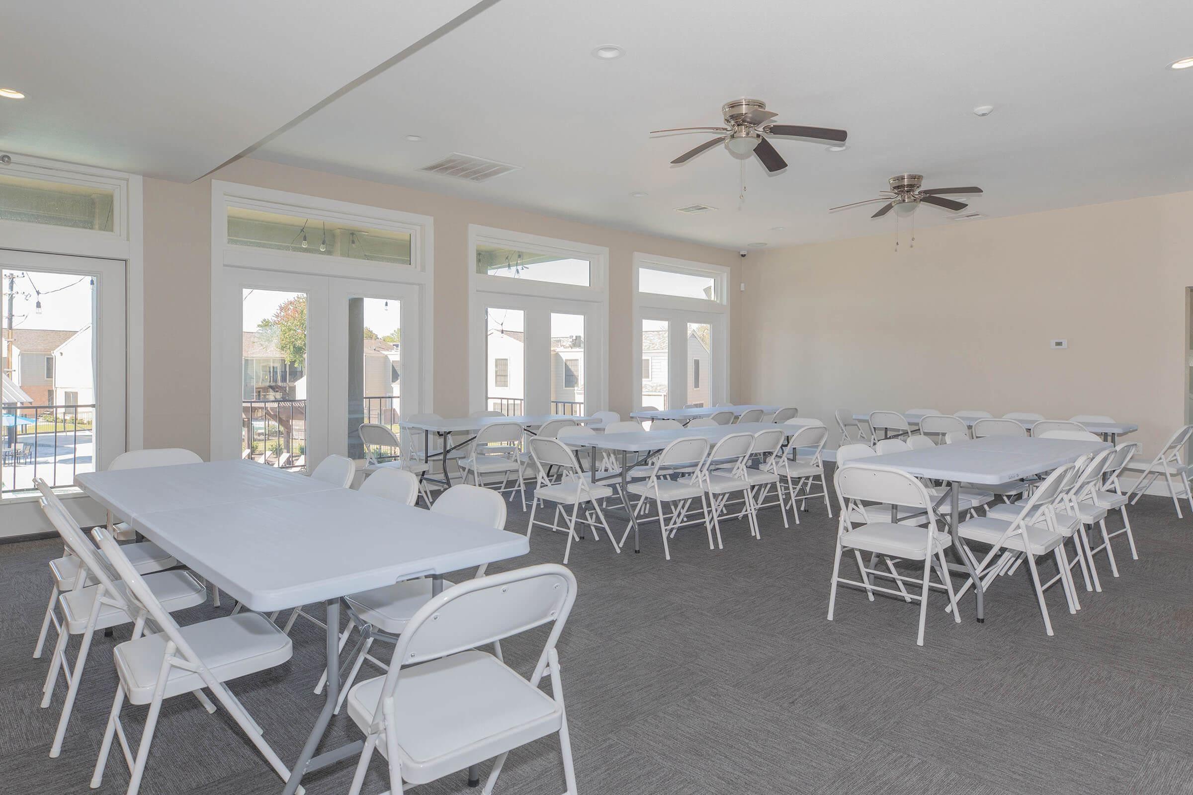An empty meeting room featuring multiple rectangular tables surrounded by white folding chairs. The space has large windows that let in natural light, ceiling fans, and a neutral color scheme with carpeted flooring. The room is ready for an event or gathering.