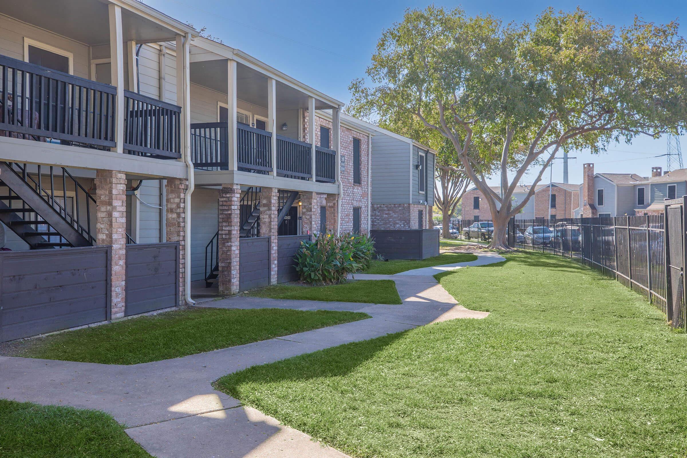 View of a landscaped pathway winding through a well-maintained apartment complex. The scene features two-story buildings with balconies, surrounded by green grass and plants, under a clear blue sky. Fences and trees add to the aesthetic appeal of the outdoor area.