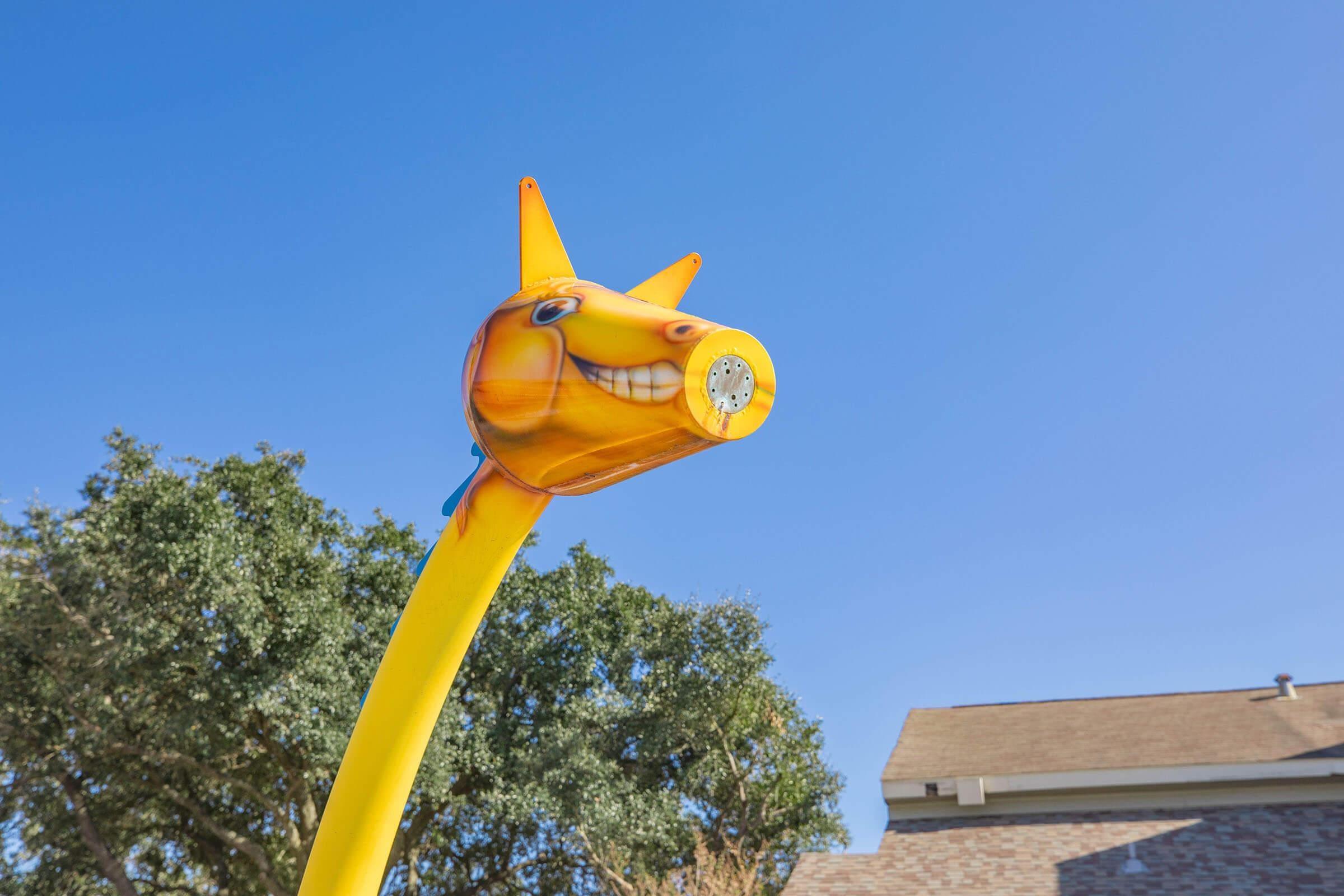 A bright yellow inflatable figure resembling a cartoon horse with a big smile, positioned against a clear blue sky, with green trees and a house in the background. The figure's head is tilted slightly upward, giving it a playful appearance.
