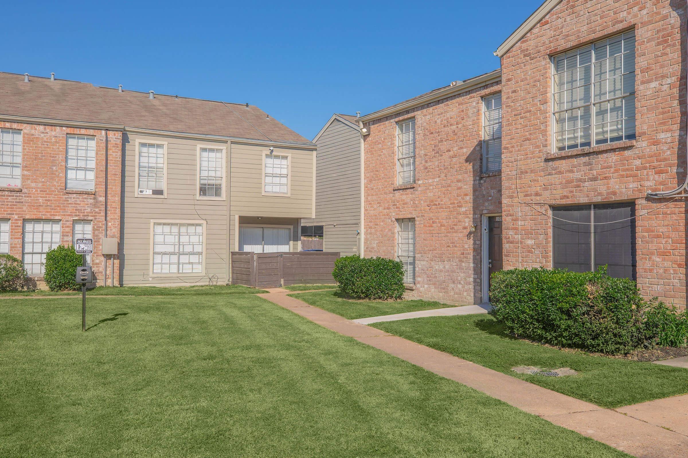 A well-maintained residential area featuring two brick apartment buildings with large windows, surrounded by neatly trimmed lawns and pathways. The sky is clear and blue, creating a bright and welcoming atmosphere.