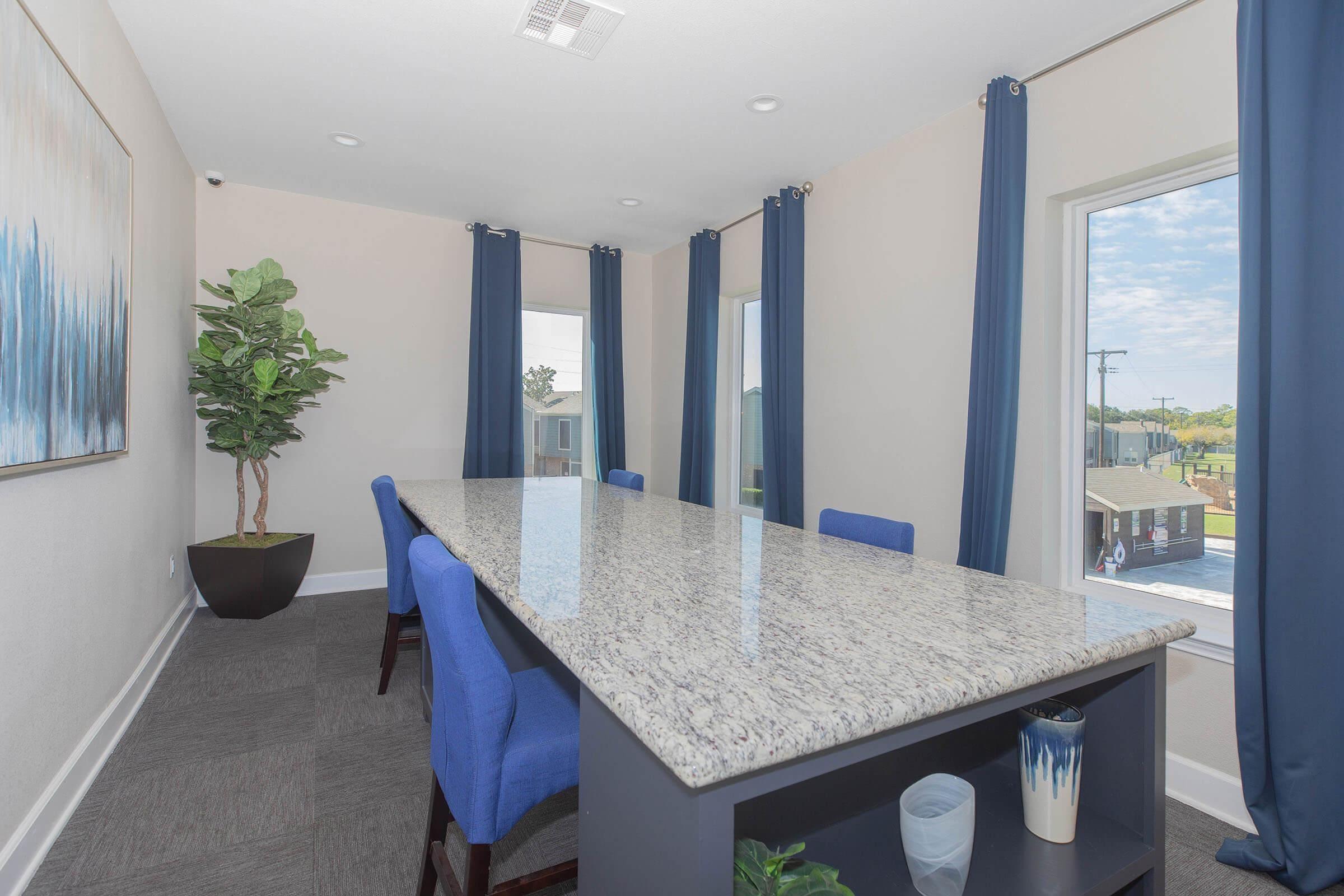 A modern meeting room featuring a long granite table surrounded by blue upholstered chairs. Large windows with blue curtains let in natural light, and a potted plant adds a touch of greenery. The walls are painted in neutral tones, creating a bright and inviting atmosphere.