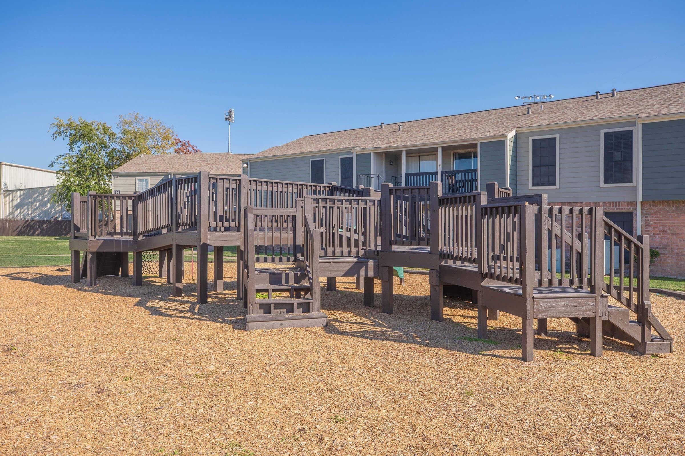 A wooden playground structure with multiple levels, ramps, and steps, surrounded by a layer of wood chips. In the background, there are two buildings with green siding and a clear blue sky. The area is well-maintained and designed for children's play.