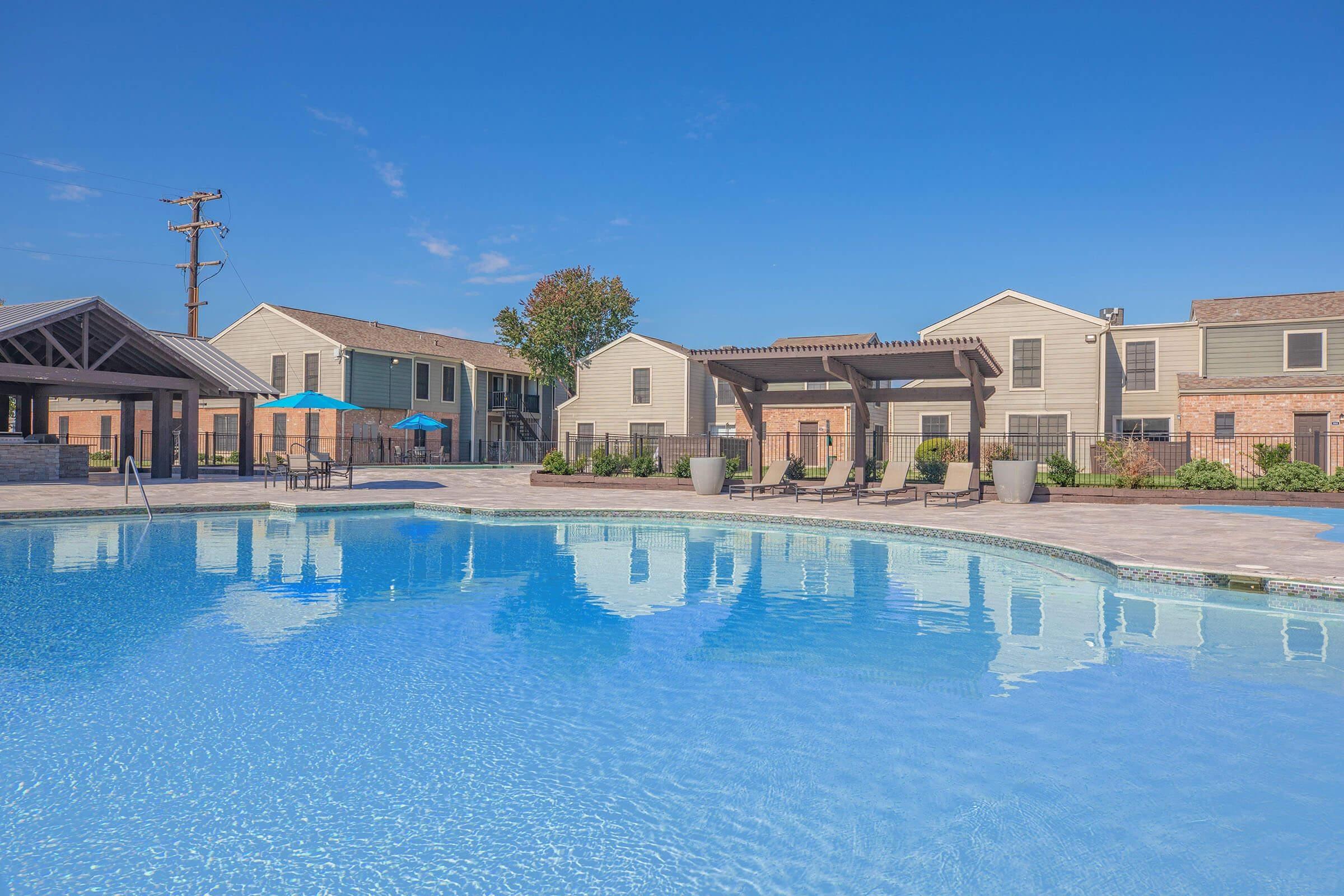 A clear blue swimming pool surrounded by well-maintained landscaping, lounge chairs, and umbrellas. In the background, there are modern apartment buildings and a shaded seating area under a pergola. The sky is bright and sunny, creating a cheerful atmosphere.