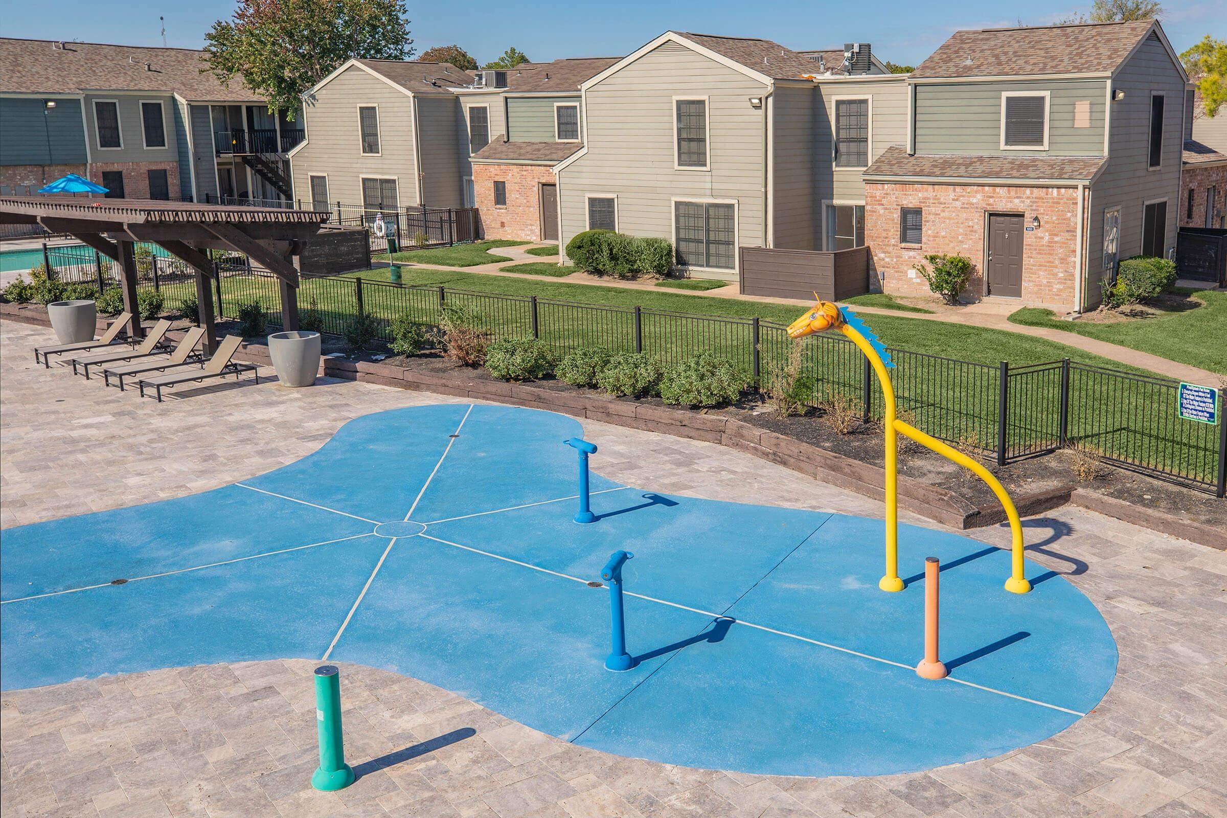 A colorful splash pad featuring a yellow giraffe water feature, surrounded by blue and orange spray nozzles. In the background, there are residential buildings with well-kept lawns and a pool area. Sunlight illuminates the scene, creating a vibrant, inviting atmosphere for families.