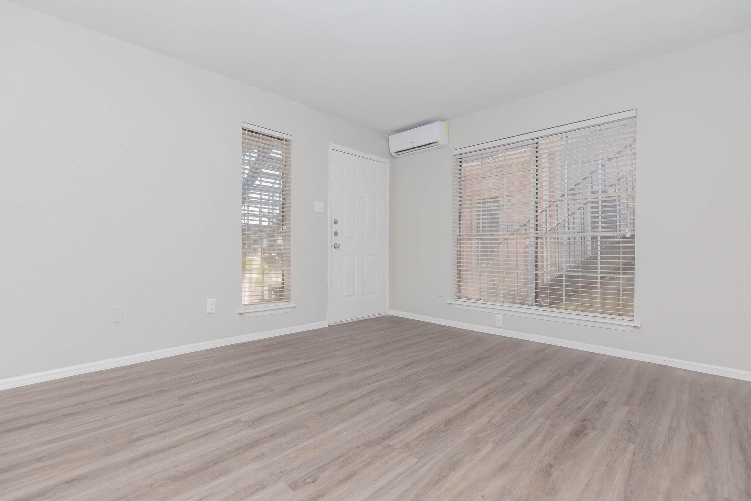 Empty room with light-colored walls and wood-like flooring. There are two windows with blinds, one on the left and one on the right side of the room, allowing natural light. A door is located on the left wall, and an air conditioning unit is mounted above the window on the right.