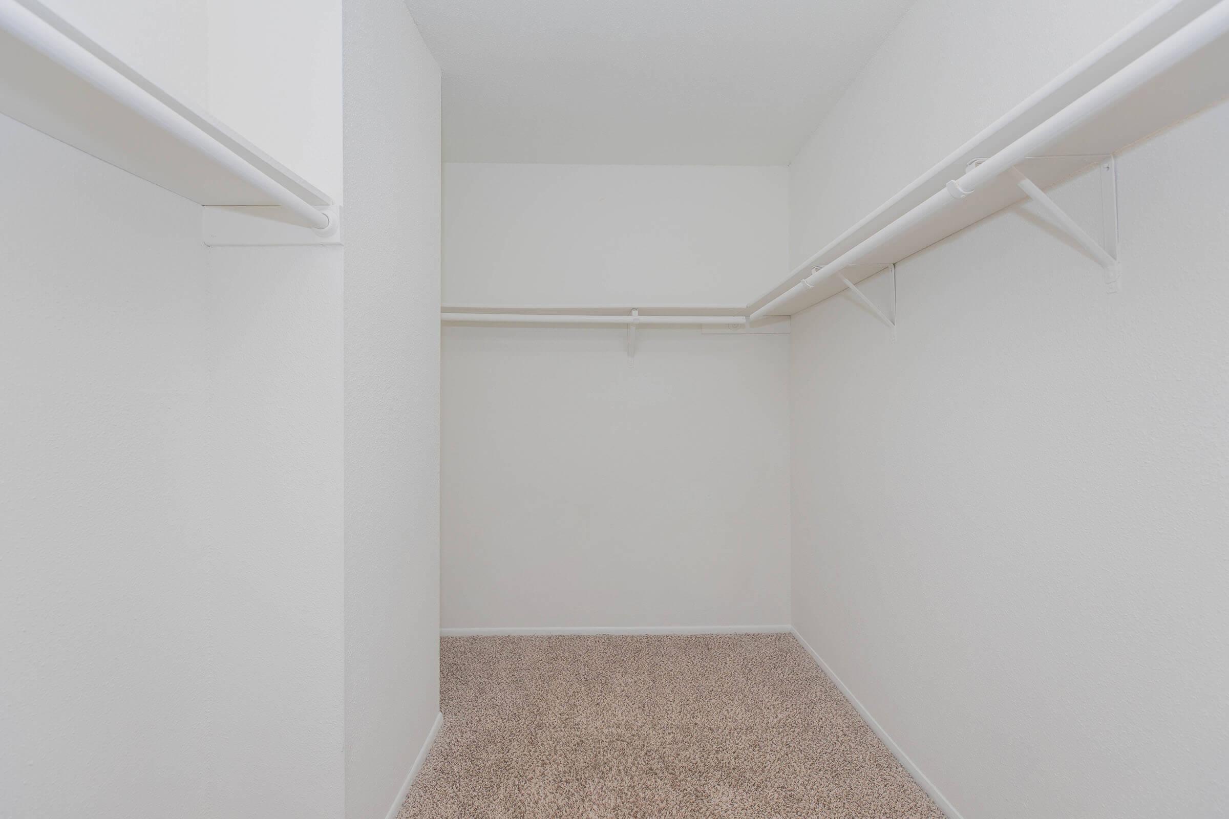 Empty walk-in closet featuring white walls, light carpet, and multiple white hanging rods on either side, providing ample space for storage or organization. The room has a clean and minimalist look, emphasizing the open space available.