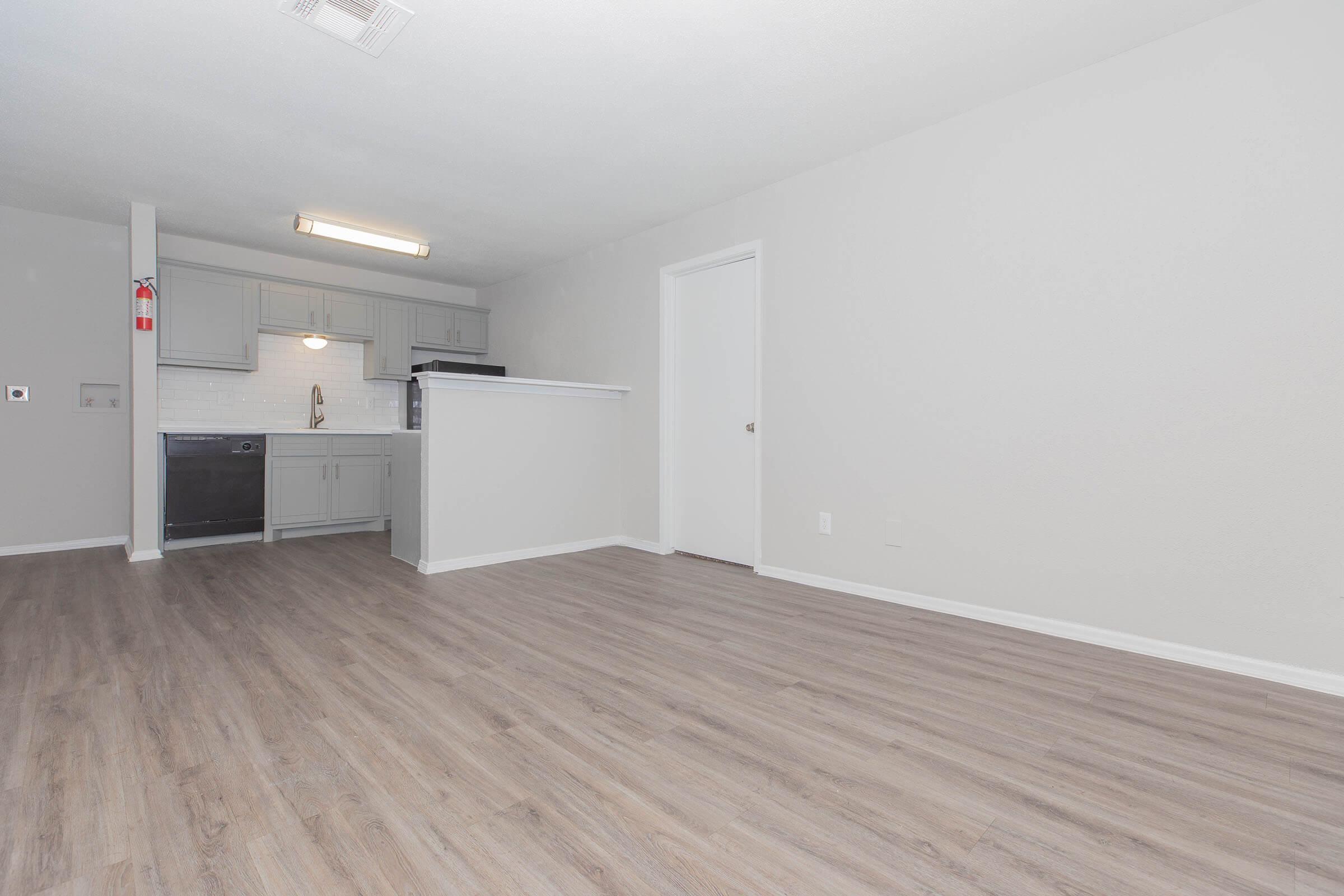 A spacious and modern kitchen area with grey cabinetry, stainless steel appliances, and white tiled backsplash. The floor is light brown laminate, and the walls are painted in neutral tones. There is a doorway leading to another room, and the space is well-lit with an overhead light fixture.