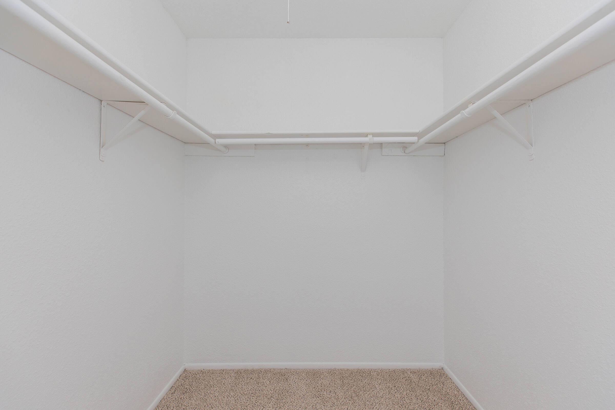 Empty walk-in closet with white walls and two hanging rods. The flooring is covered in beige carpet, creating a spacious and clean appearance.