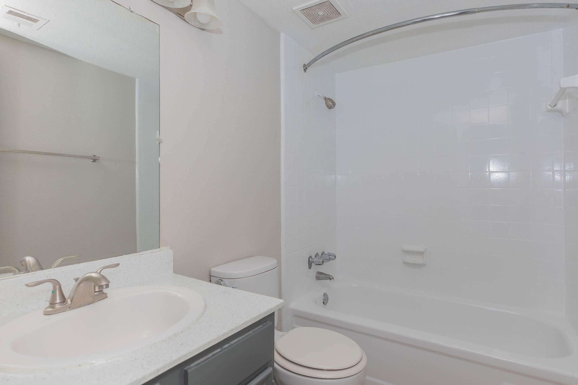 A clean, modern bathroom featuring a bathtub with a curved shower rod, a toilet, and a sink with a gray cabinet. The walls are painted white, and there's a large mirror above the sink. Natural light illuminates the space, creating a fresh and inviting atmosphere.