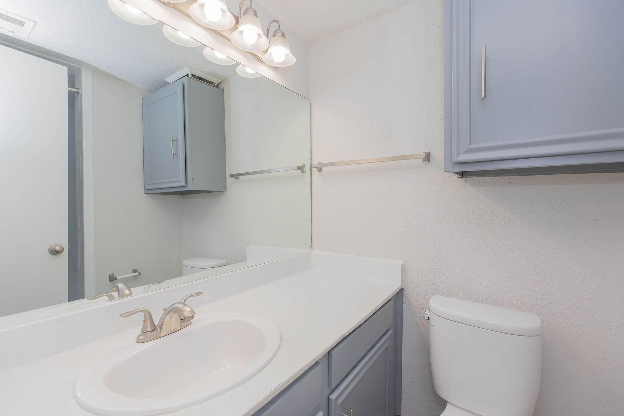 A clean, modern bathroom featuring a white countertop with a sink, a framed mirror above, soft blue cabinets, and a toilet. There are five light fixtures above the mirror, and a shower area is visible in the background, creating a bright and inviting space.