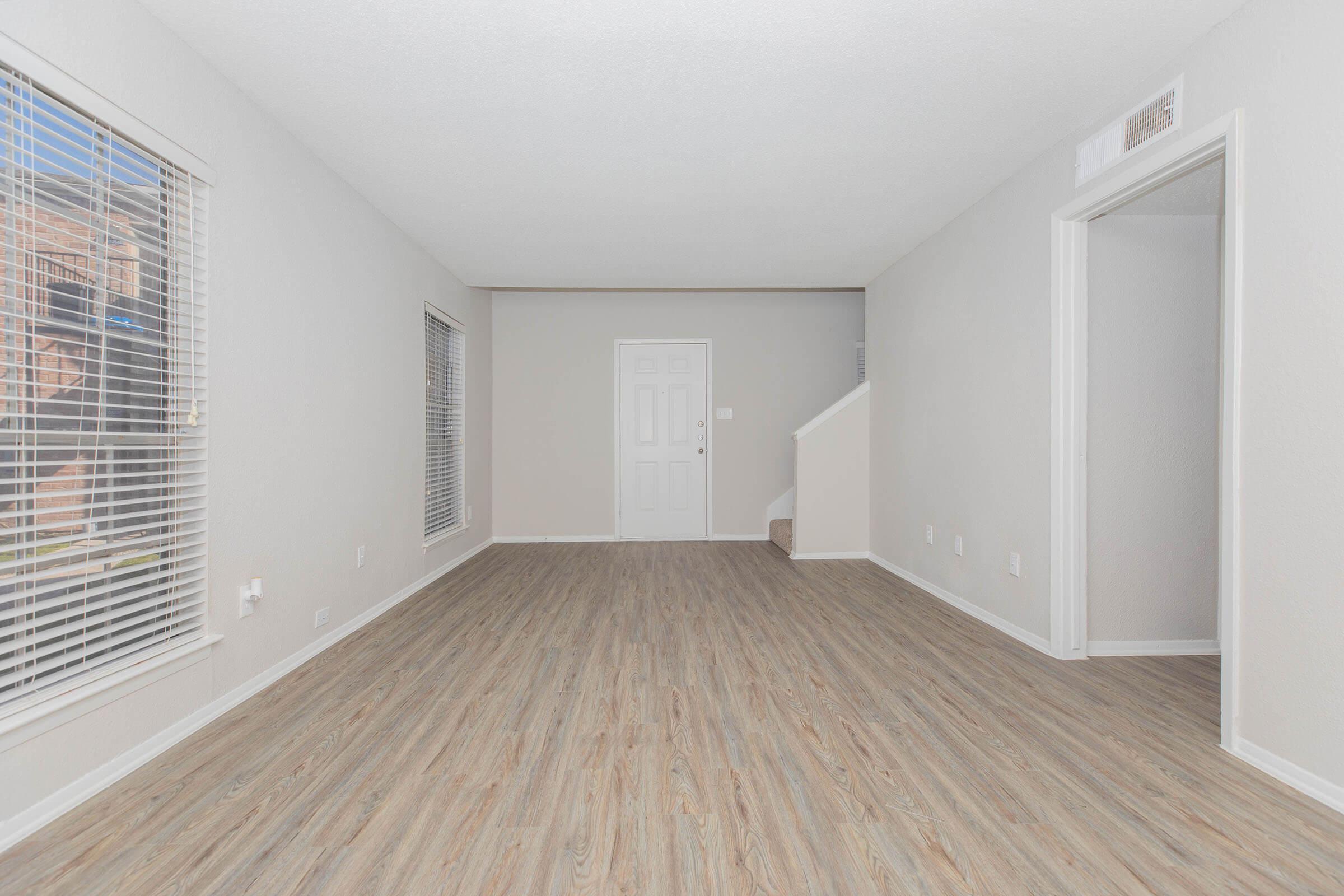 A spacious, empty living room with light gray walls and light wood laminate flooring. Two windows with blinds provide natural light. A closed door is visible on one wall, and a staircase is seen in the background. The room appears clean and ready for furnishings.
