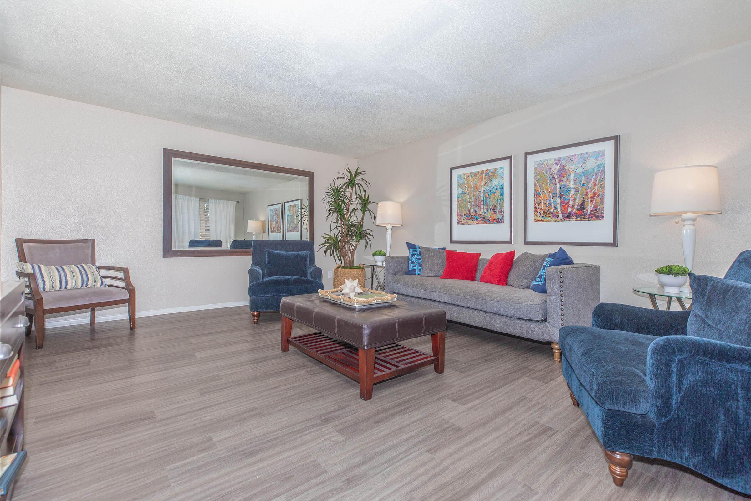 A spacious living room featuring a gray sofa with red and blue accent pillows, two blue armchairs, a wooden coffee table with decorative items, and a large mirror on the wall. The room has light-colored walls and wooden flooring, with a plant in the corner and framed colorful artwork on display.