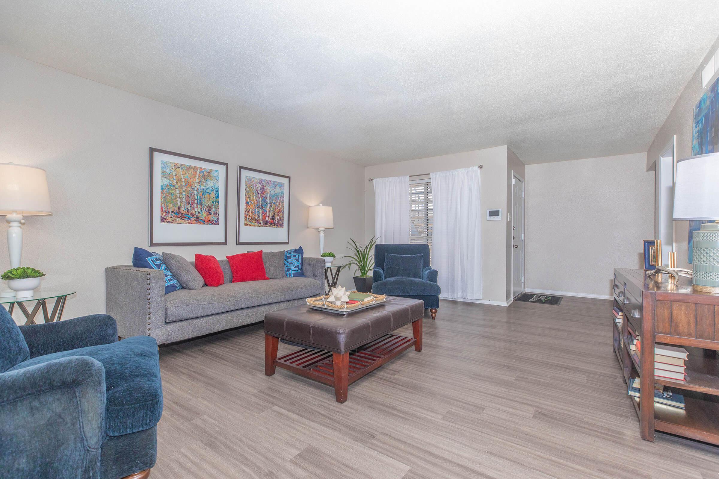A cozy living room featuring a gray couch with red and blue accent pillows, two blue armchairs, and a wooden coffee table. There are colorful framed artworks on the wall, a lamp beside the couch, and a plant in the corner. Natural light filters in through a window with white curtains, creating a warm atmosphere.