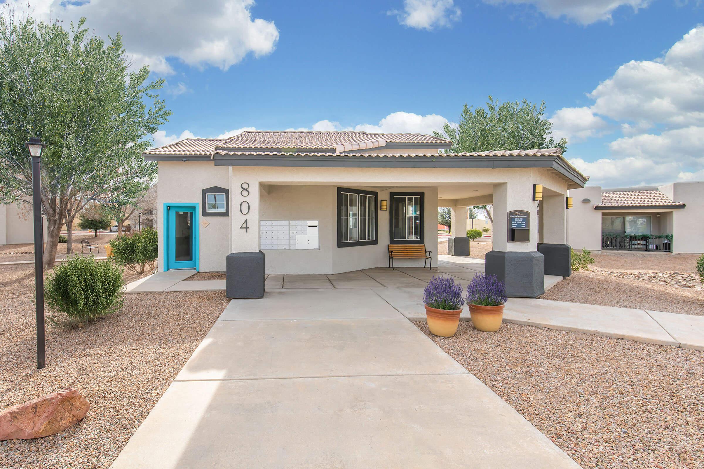 A single-story, modern home with a light-colored exterior and a tiled roof. The front yard features gravel landscaping, two decorative pots with lavender plants, and a walkway leading to the entrance. Nearby are low shrubs and a lamp post, with a clear blue sky and clouds in the background.