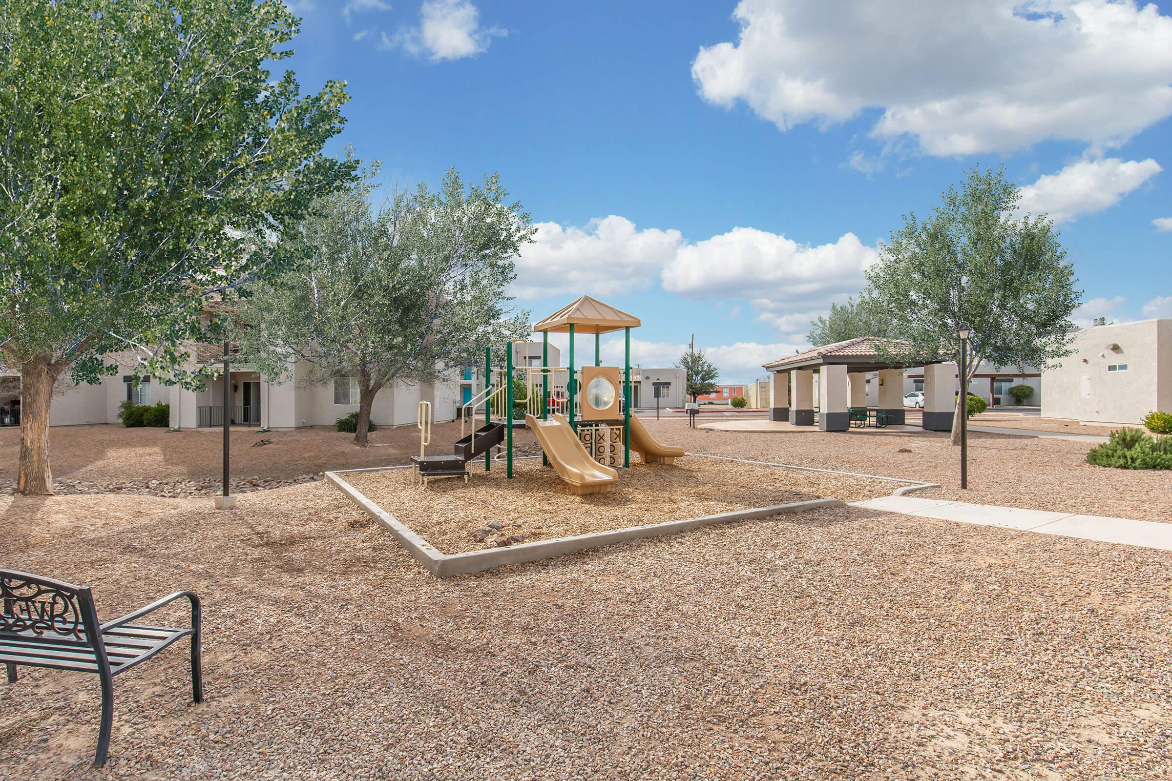 A playground featuring a slide and climbing structure, surrounded by gravel and grassy areas. In the background, there are trees and small buildings under a cloudy sky, creating a peaceful outdoor setting. A bench is visible in the foreground.