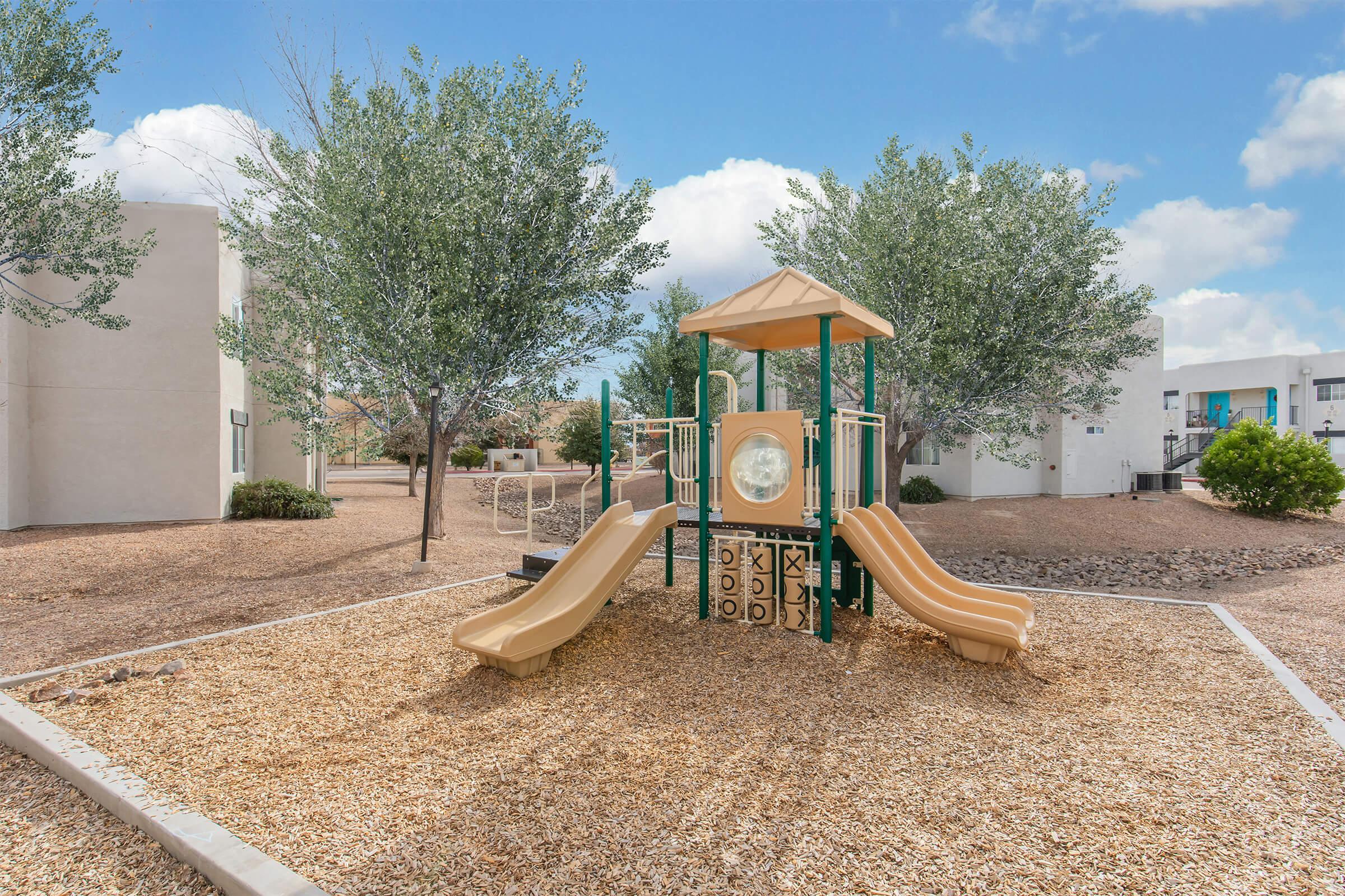Playground with two slides, a small climbing structure, and a circular window, surrounded by a gravel area and trees. Residential buildings are visible in the background under a blue sky with fluffy clouds.