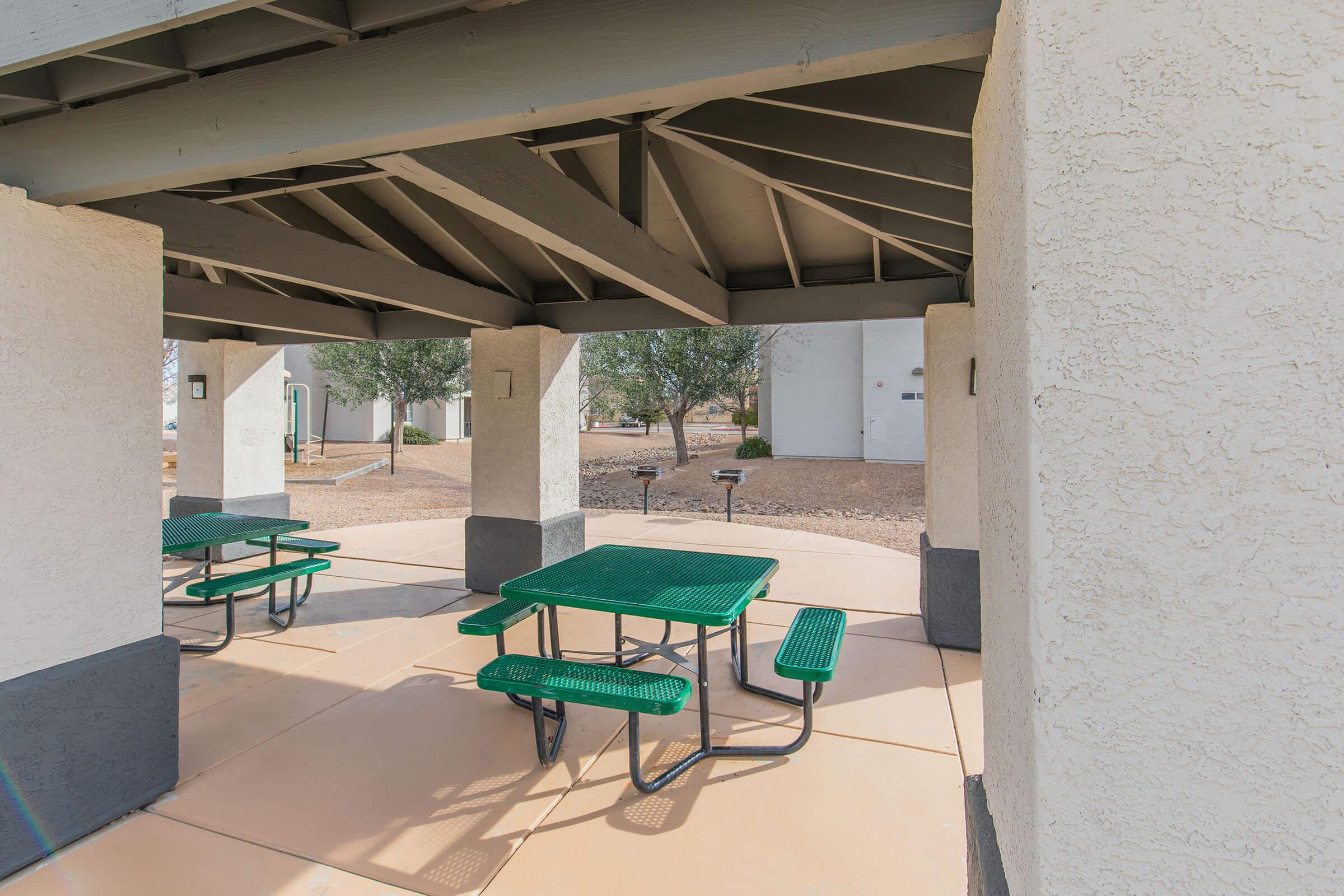 Covered outdoor area with green picnic tables and benches, surrounded by a light-colored concrete structure. The space is open with a view of the surrounding ground and some trees in the background.