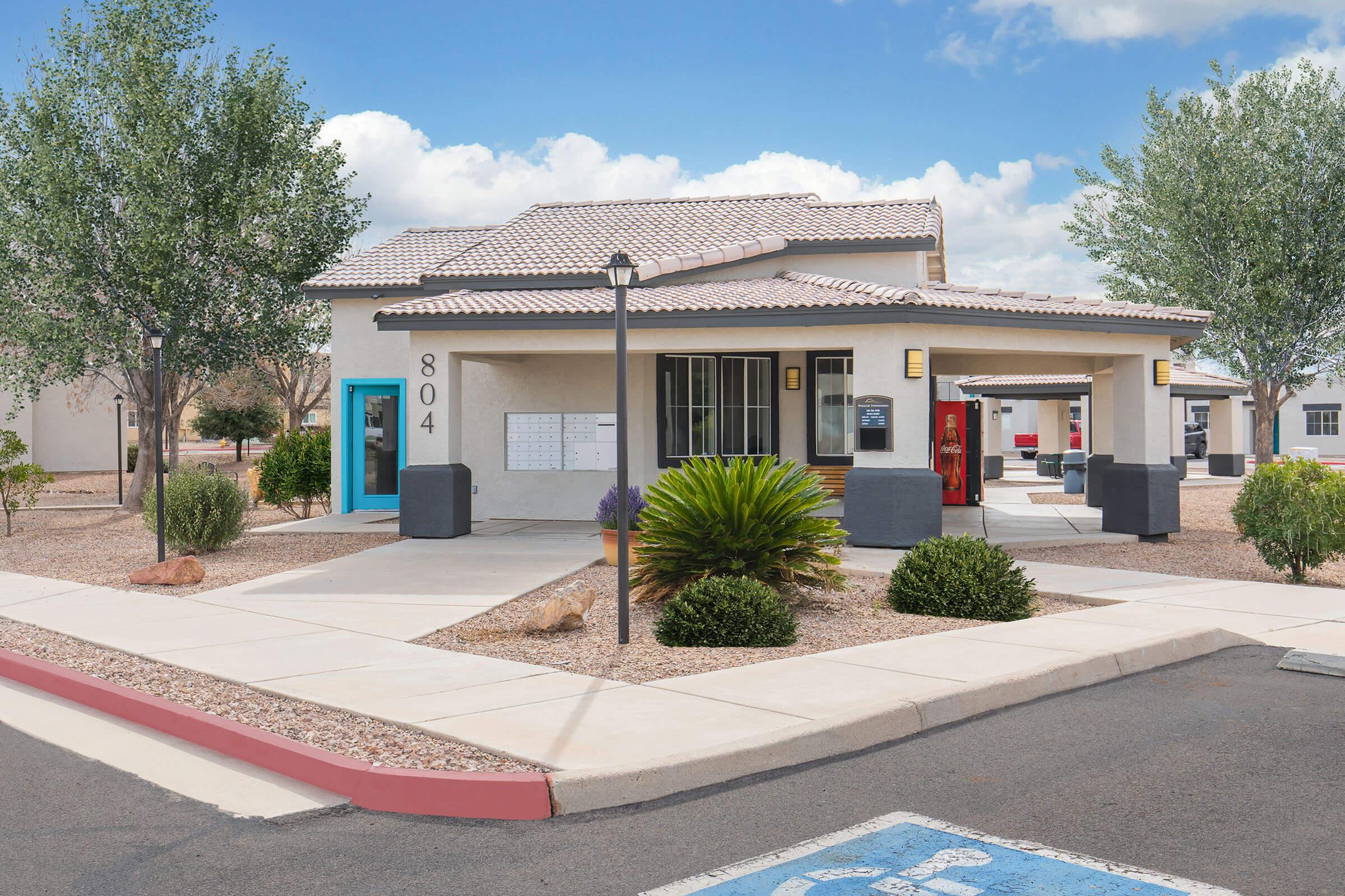 A single-story building with a beige exterior and a tiled roof, featuring blue and black doors. It has landscaped areas with shrubs and small trees, a paved pathway, and a parking space marked for accessibility. The scene is set against a partly cloudy sky.