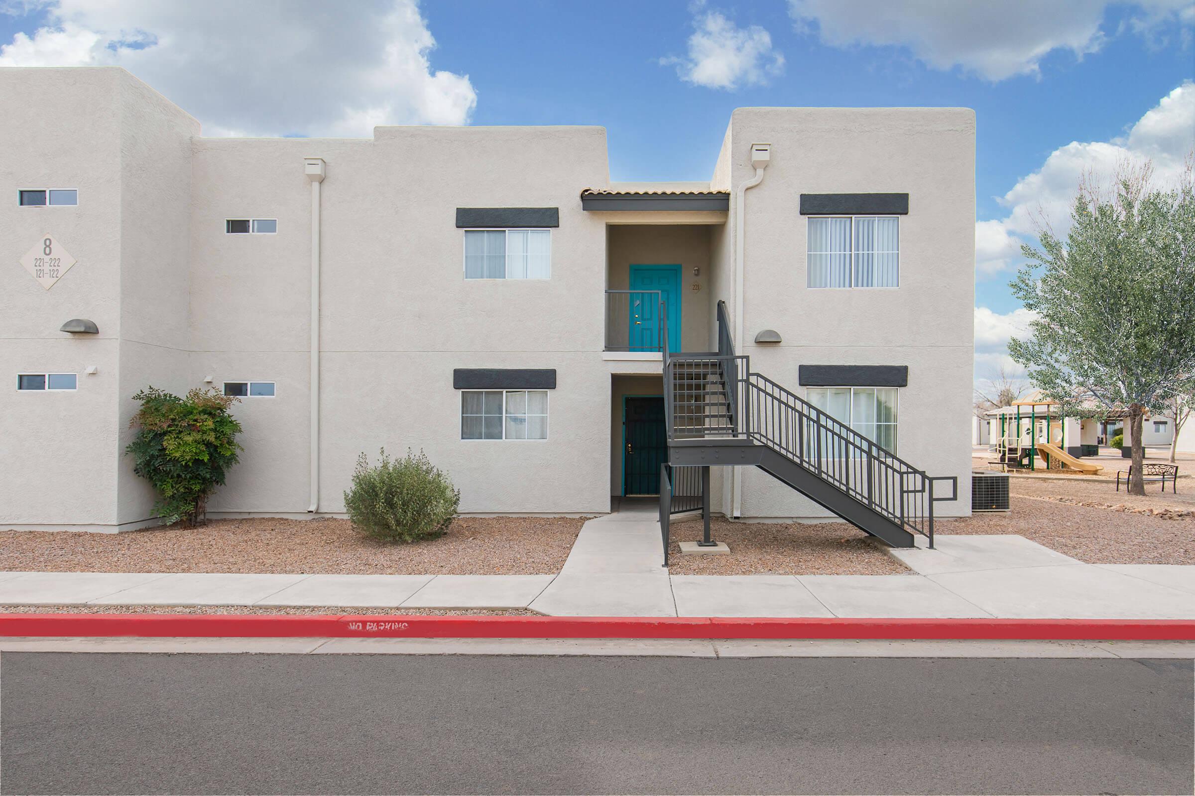 A beige two-story apartment building with a turquoise door, staircase, and walkway. The surrounding area features desert landscaping with bushes and small trees, under a partly cloudy blue sky. A playground can be seen in the background, along with additional buildings.