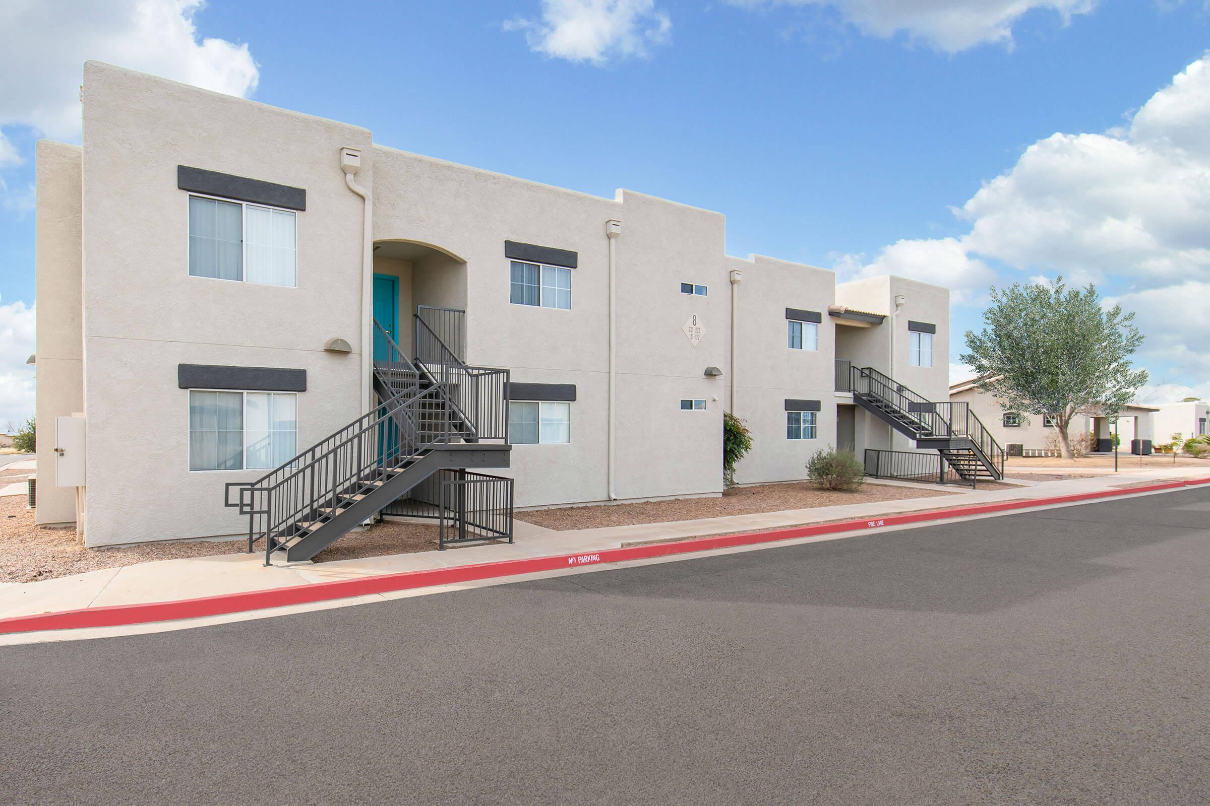 A modern, two-story apartment building with a light-colored exterior. It features multiple entrances with metal stairs leading to the second floor. There are small plants and shrubs near the building, and the area is well-maintained with a clear blue sky and scattered clouds in the background.