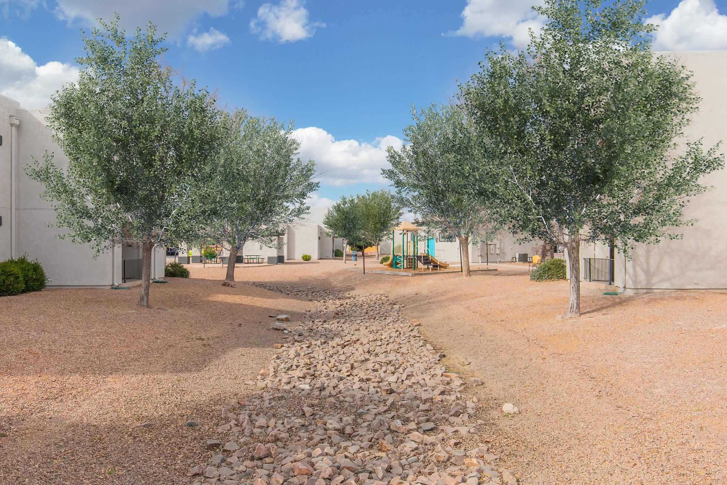 A tranquil outdoor scene featuring a pathway made of rocks lined with green trees. The background includes low buildings and a playground under a blue sky with clouds, creating a serene atmosphere.