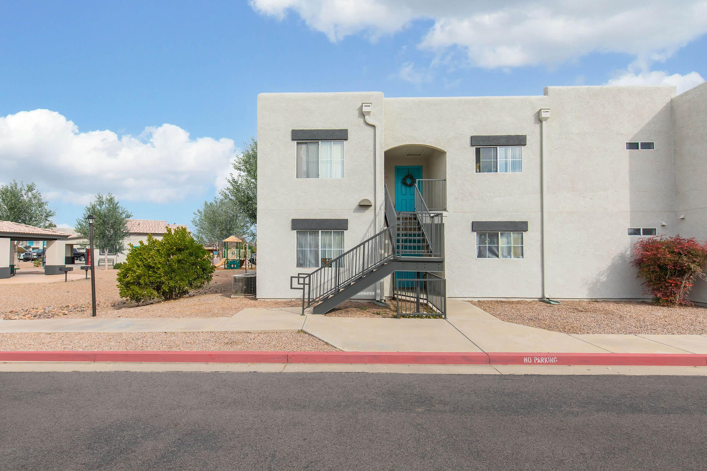 A two-story apartment building with light-colored stucco exterior and dark trim. An exterior staircase leads to the second floor, where a door is visible. The surrounding area features desert landscaping, including small trees and gravel. The sky is partly cloudy, and a "No Parking" sign is visible nearby.