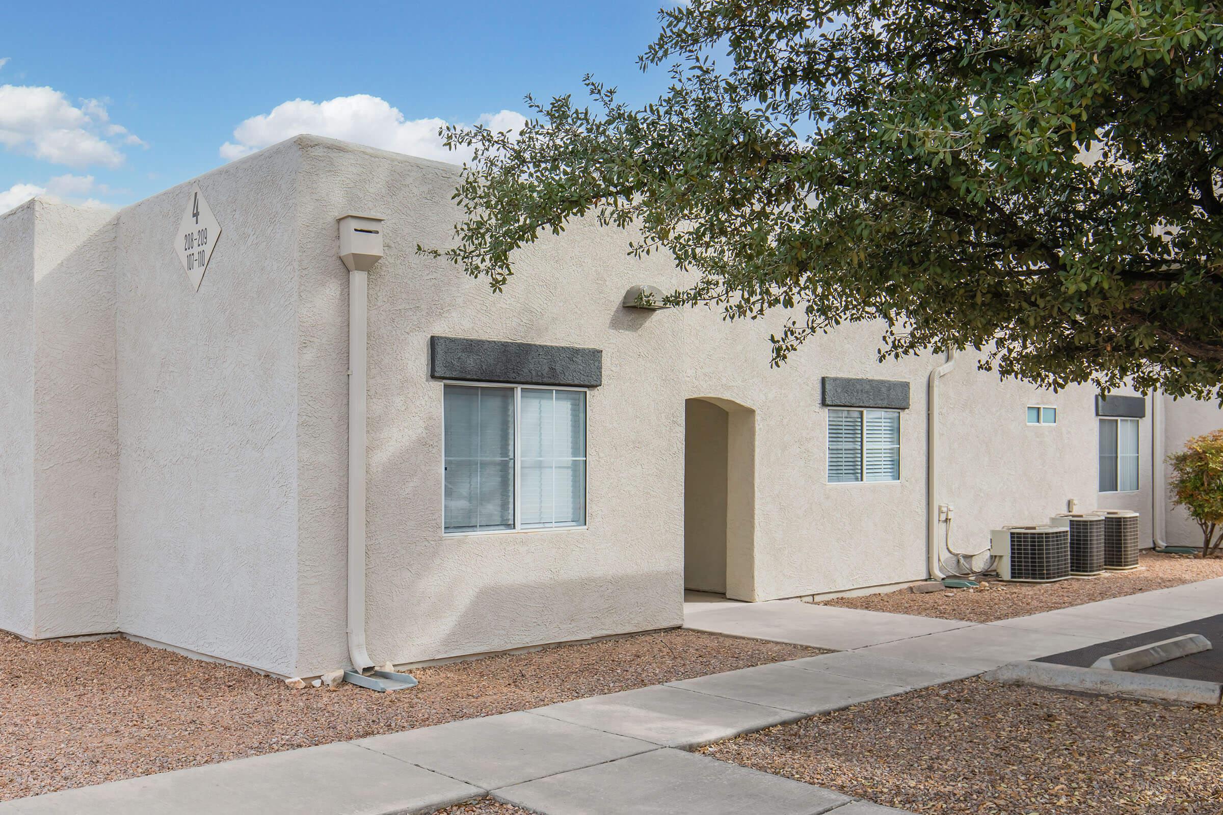 A light-colored, modern single-story apartment building with a concrete walkway leading to the entrance. The building features two windows with dark trim and a small tree nearby. There are air conditioning units visible on the side, and the landscape includes small gravel. The sky is partly cloudy.