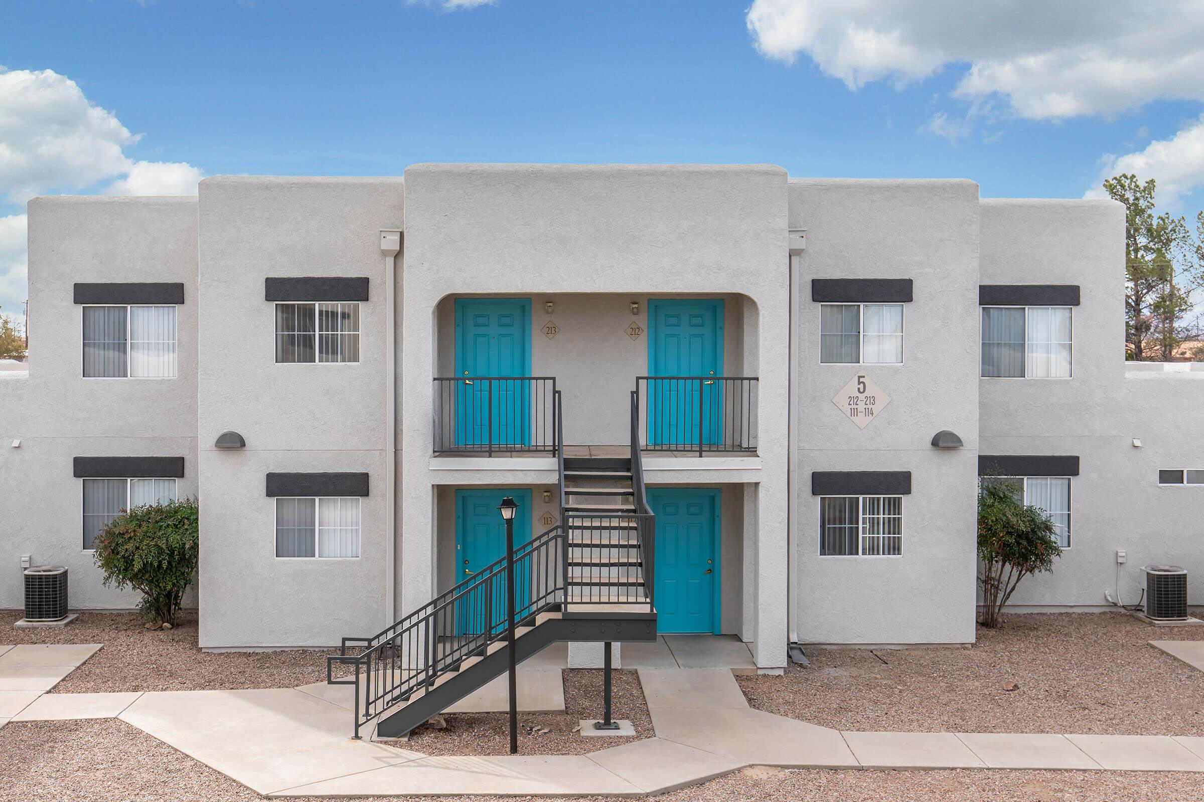 Two-story apartment building with a light gray exterior and turquoise doors. The front features a central staircase leading to the second floor. There are several windows adorned with white curtains on each side. The surrounding area includes gravel landscaping and small shrubs. The sky is partly cloudy.