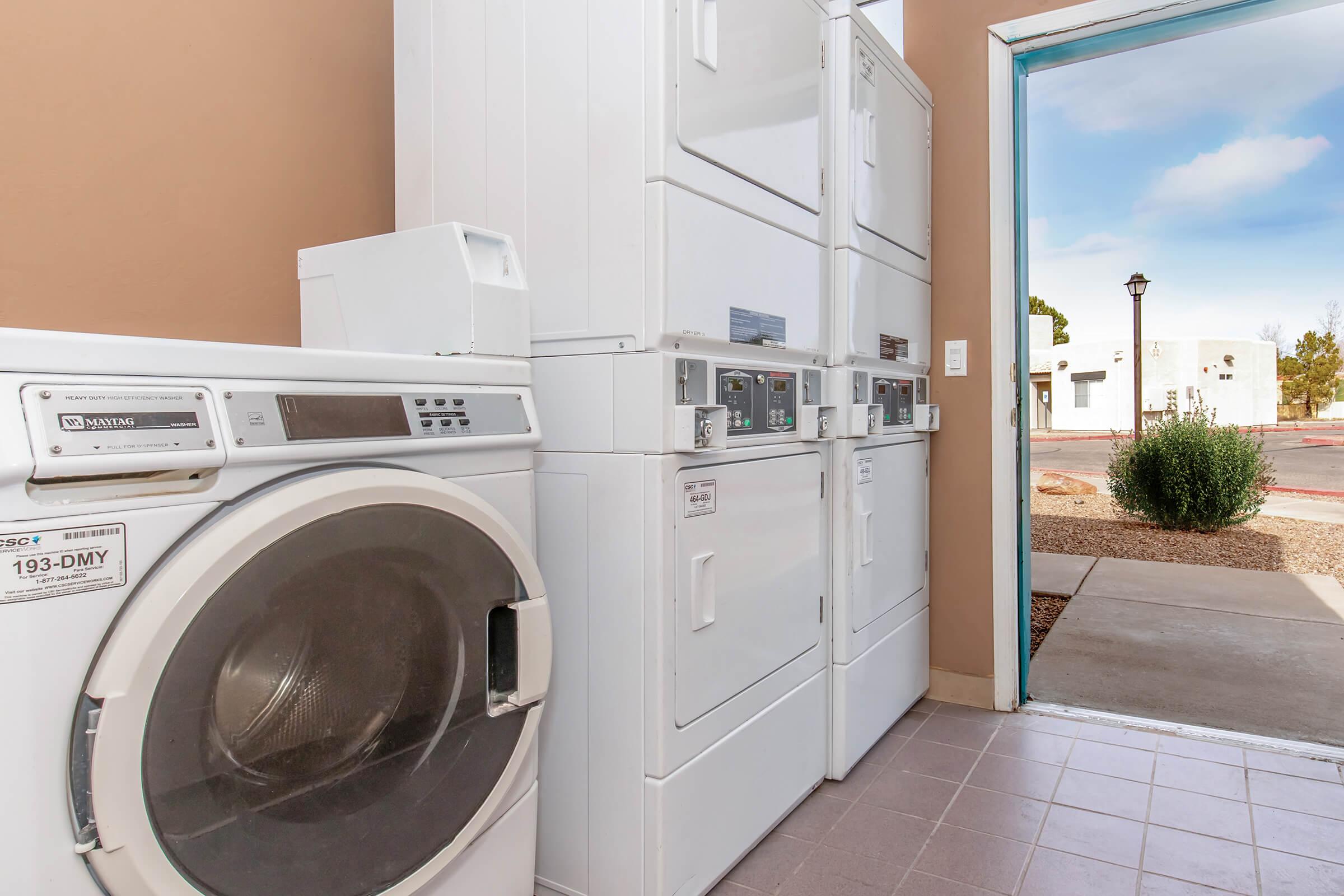 A laundry room featuring stacked washer and dryer units, with a door leading to an outdoor area. The floor is tiled, and there's a window providing natural light. The setting is clean and organized, with a few shrubs visible outside.