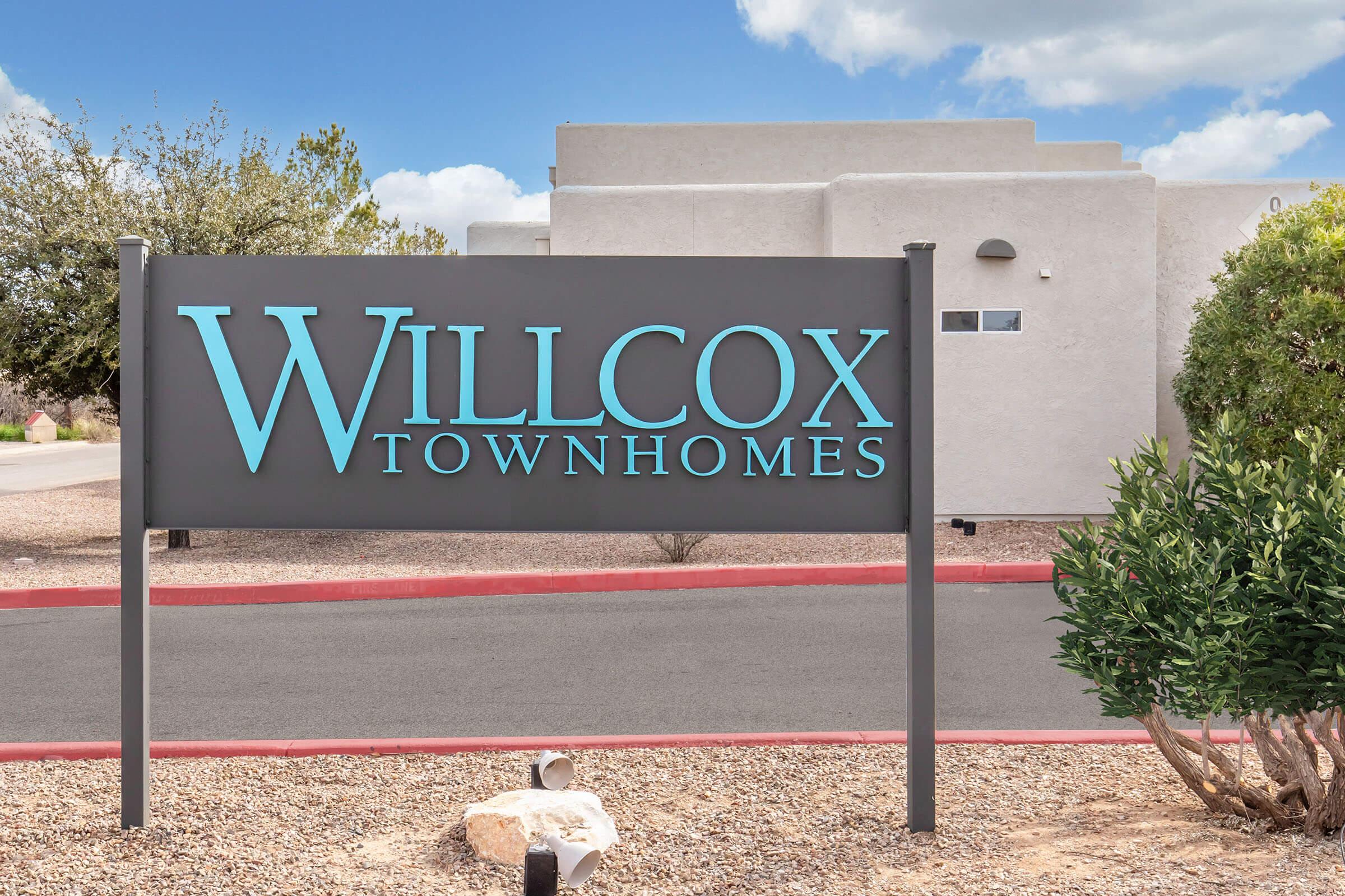 Sign for Willcox Townhomes featuring a modern design with bold blue and white lettering against a simple gray background. Surrounded by desert landscaping and a clear blue sky, it indicates the location of the townhomes.