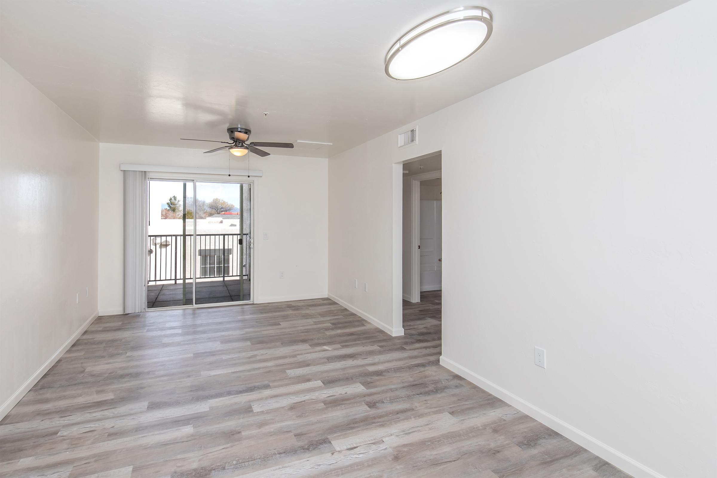 Empty room with light-colored walls and laminate flooring. A ceiling fan is visible above, along with a modern circular light fixture. There is a sliding glass door leading to a balcony, and another doorway on the right side leading to a separate space or room.