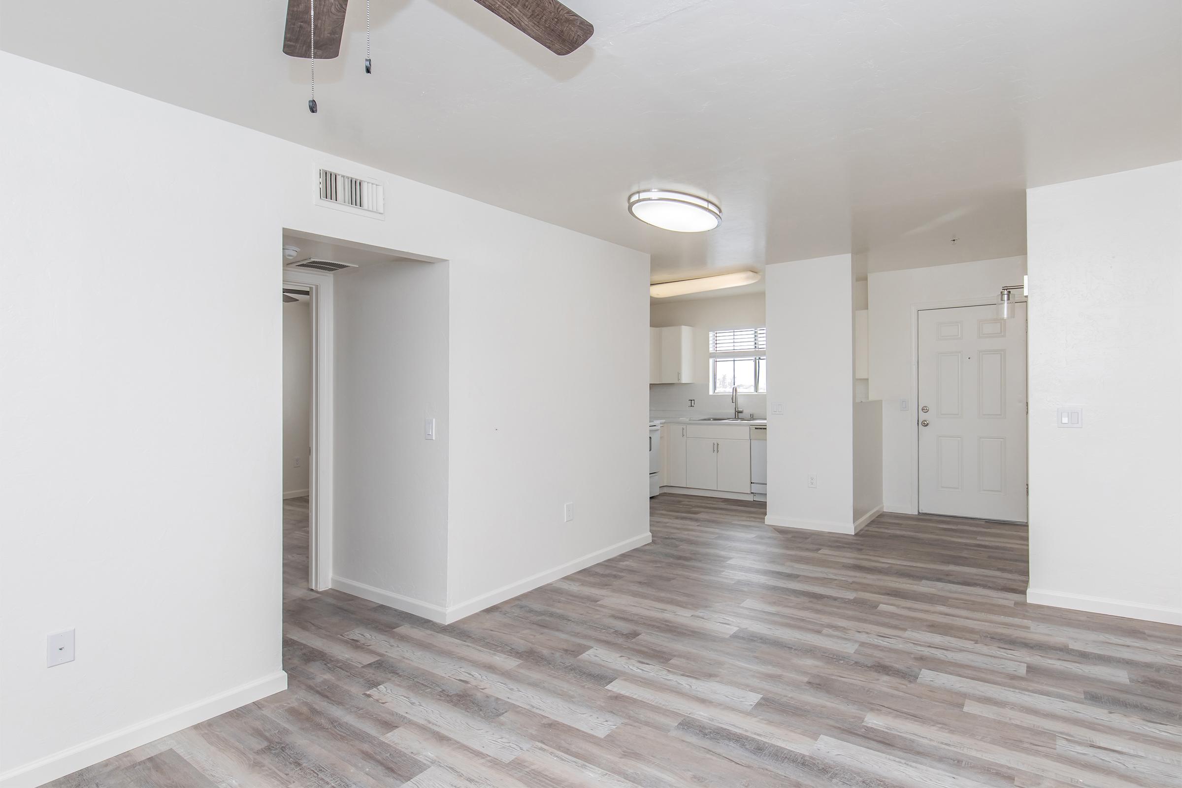 Bright and spacious interior view of an apartment featuring an open layout. The living area with new wood-like flooring leads to a kitchen with white cabinetry. A ceiling fan hangs above, and a door provides access to the outside. Soft natural light fills the space through windows.