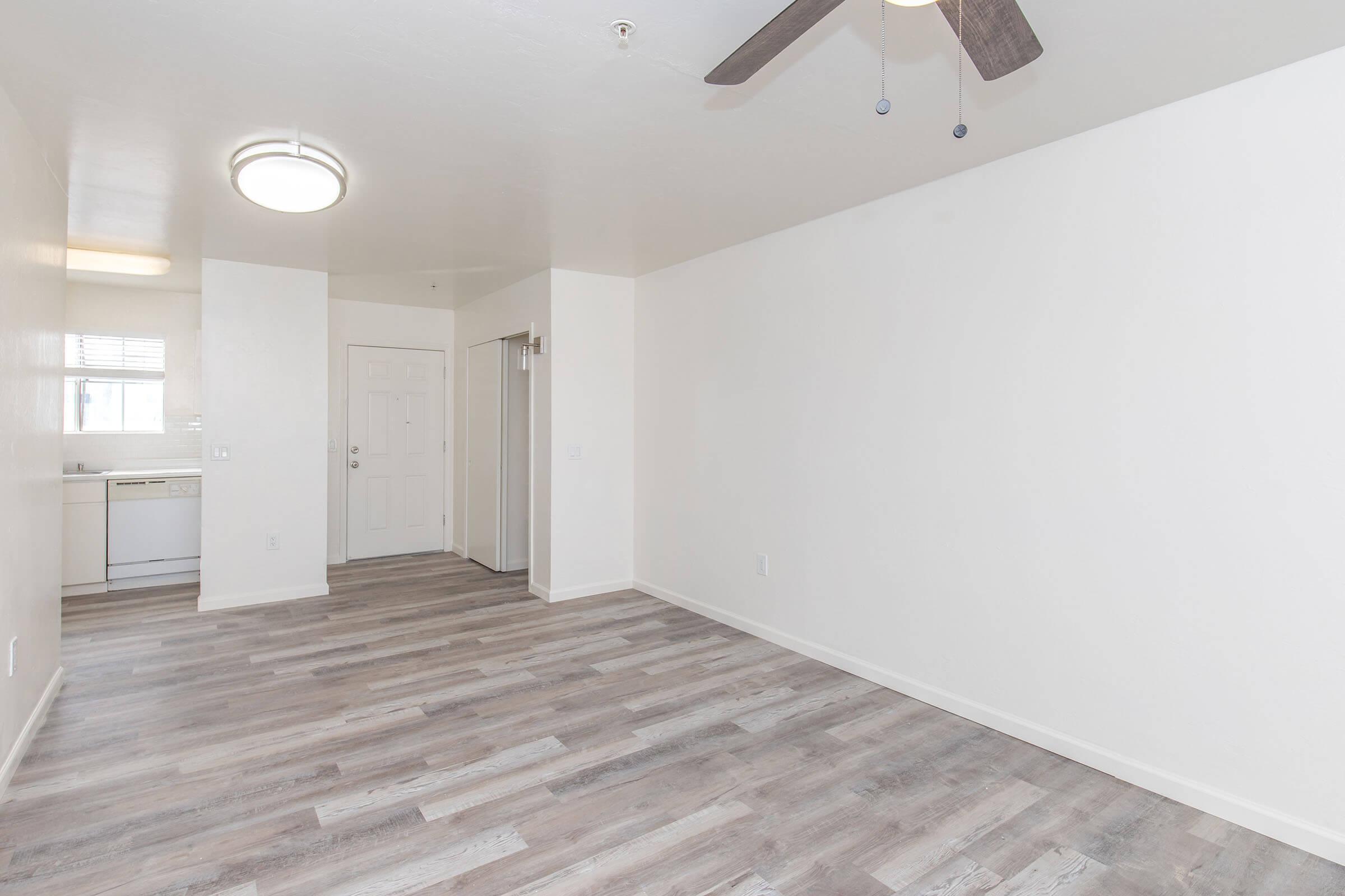 Interior view of an empty room featuring light-colored walls and wooden flooring. A ceiling fan hangs from the ceiling, and there is a light fixture above. The space includes an open door to a kitchen area on the left and a doorway leading to another room on the right. Natural light comes from a window.