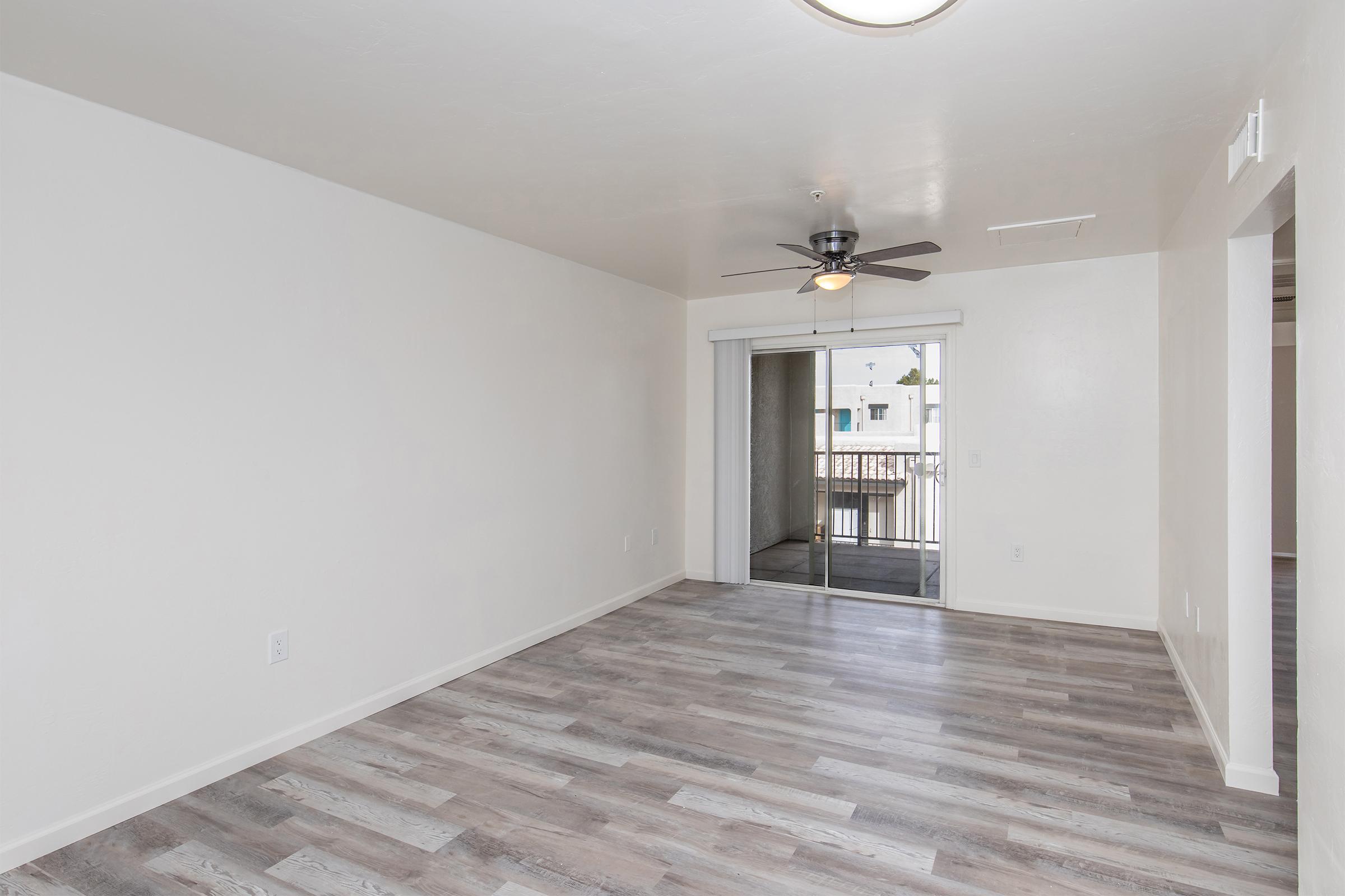A spacious, unfurnished living room featuring light-colored walls, a ceiling fan, and large sliding glass doors leading to a balcony. The floor is covered with light wood laminate, creating a bright and airy atmosphere.