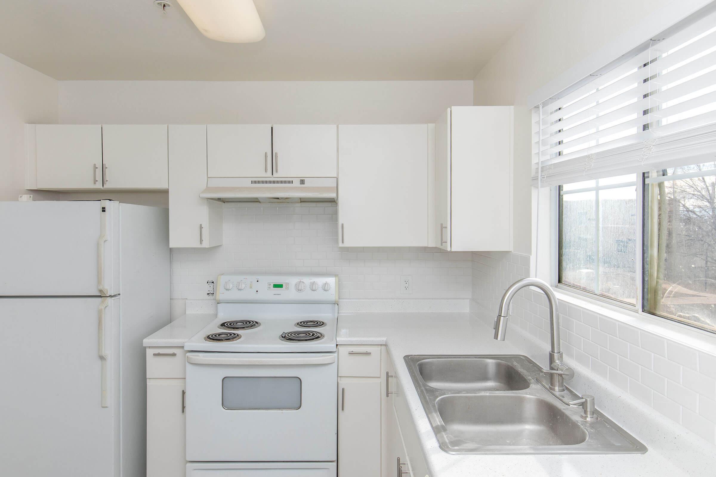 A modern kitchen with white cabinets, a white refrigerator, and a white stove with black burners. The countertop is light-colored, and there are two stainless steel sinks. A window above the sink allows natural light to enter, with a view of the outdoors. The overall design is clean and minimalist.