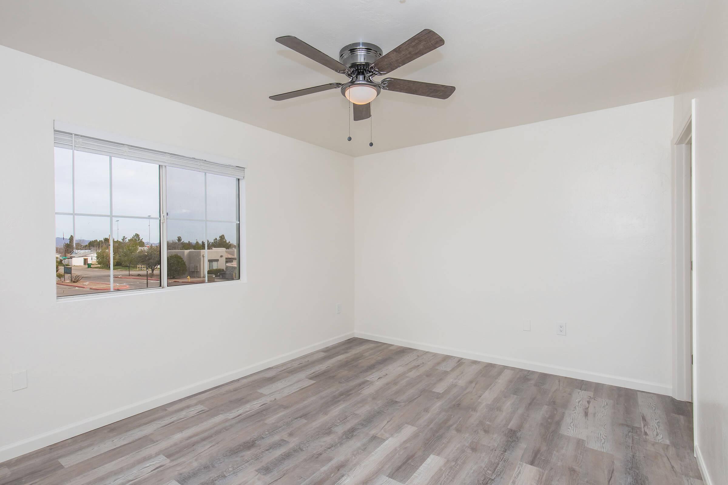 A spacious, empty room with light-colored walls and a ceiling fan. The floor is covered in light gray laminate. A large window provides natural light and shows views of a neighborhood and palm trees outside. The overall atmosphere is bright and fresh, suitable for various uses.