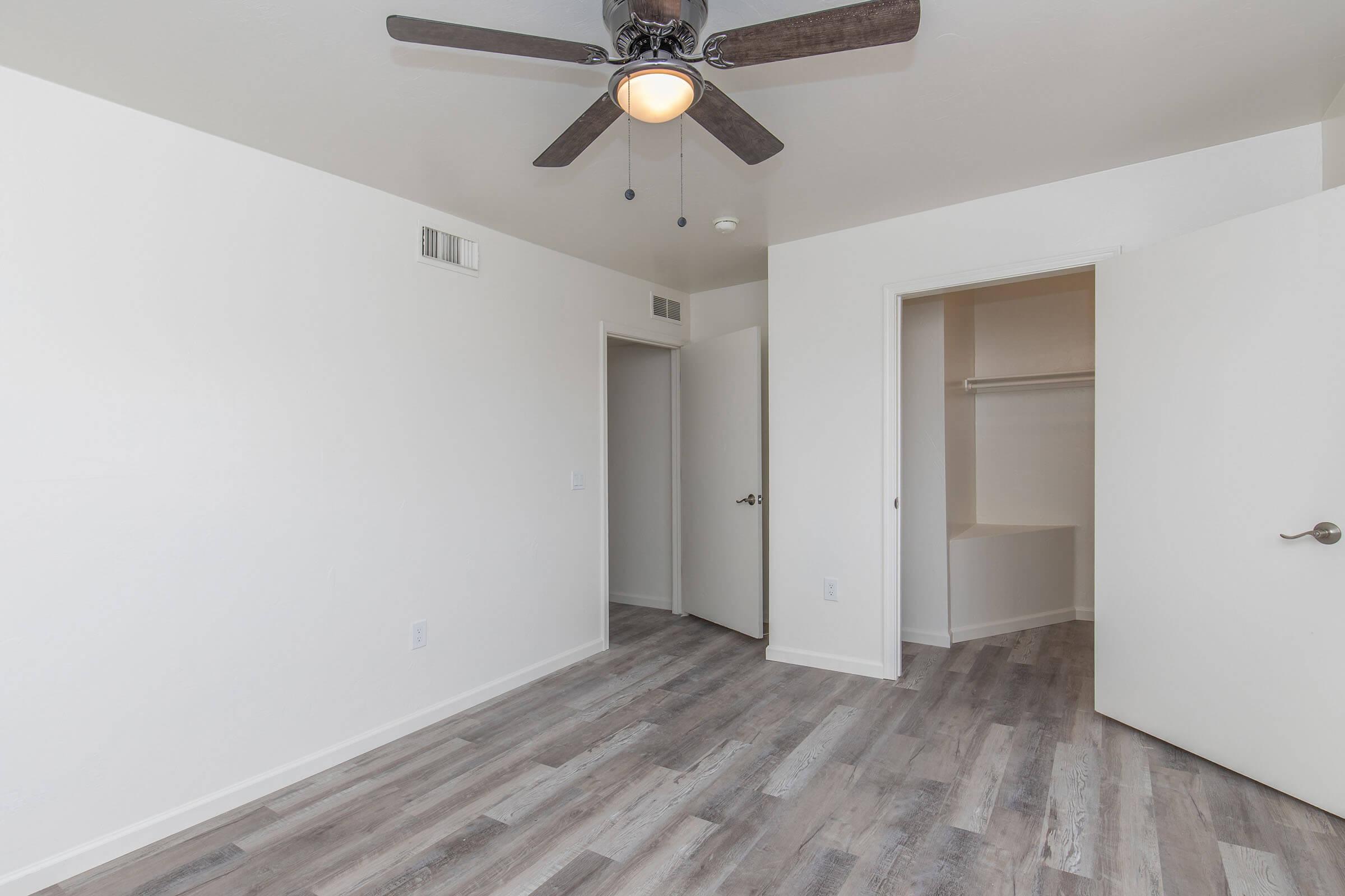 Empty room with light-colored walls and gray wood-like flooring. A ceiling fan with wooden blades hangs from the ceiling. Two doors are visible: one leads to a closet, and the other opens to an adjacent room. The space appears bright and well-lit, with a simple, modern design.
