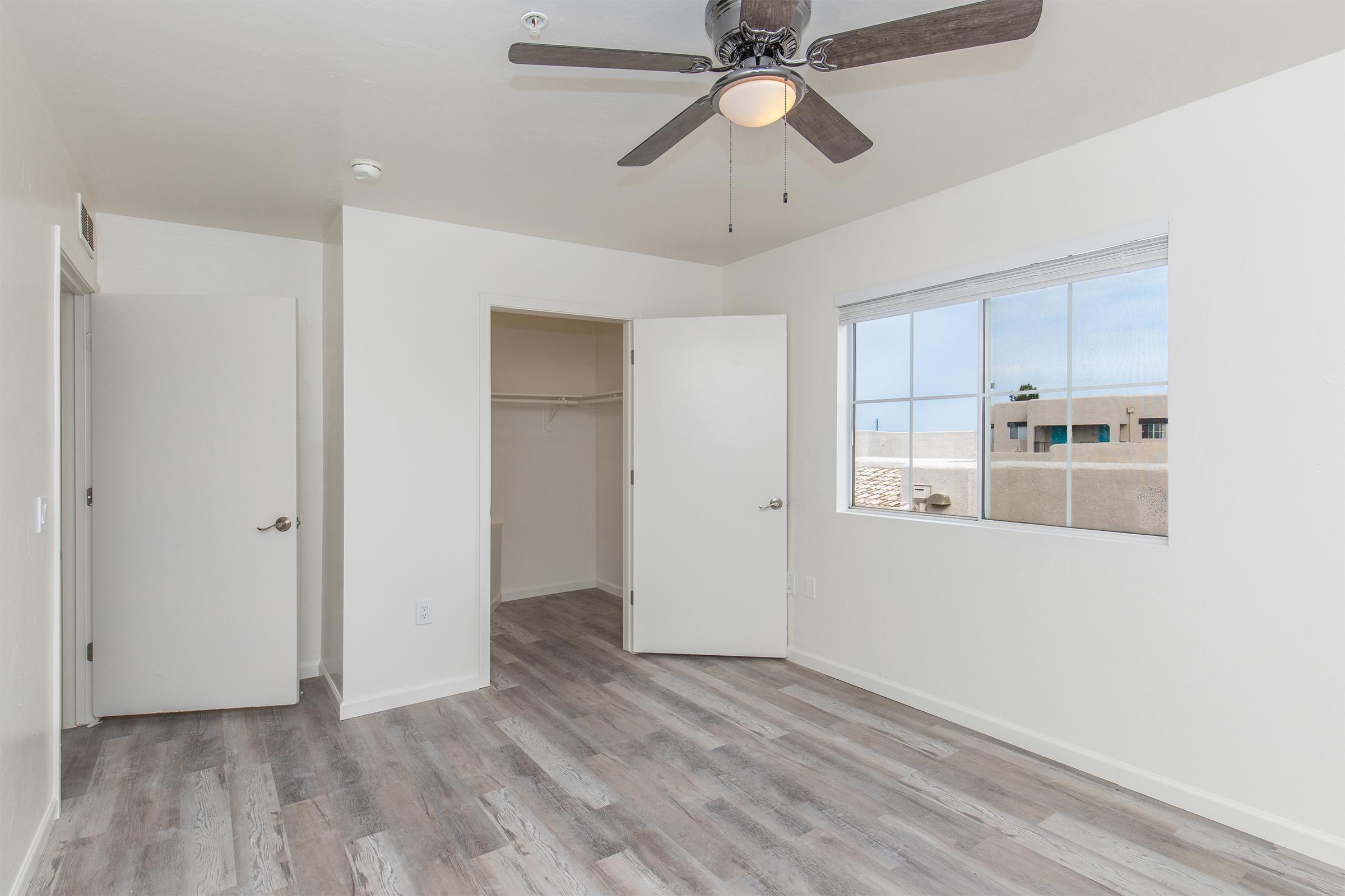 Interior of a bright, airy bedroom featuring light-colored walls and wood-like laminate flooring. One wall has a window allowing natural light, and opposite are two open doors leading to a closet and another area. A ceiling fan is mounted above, adding to the room's comfort and style.