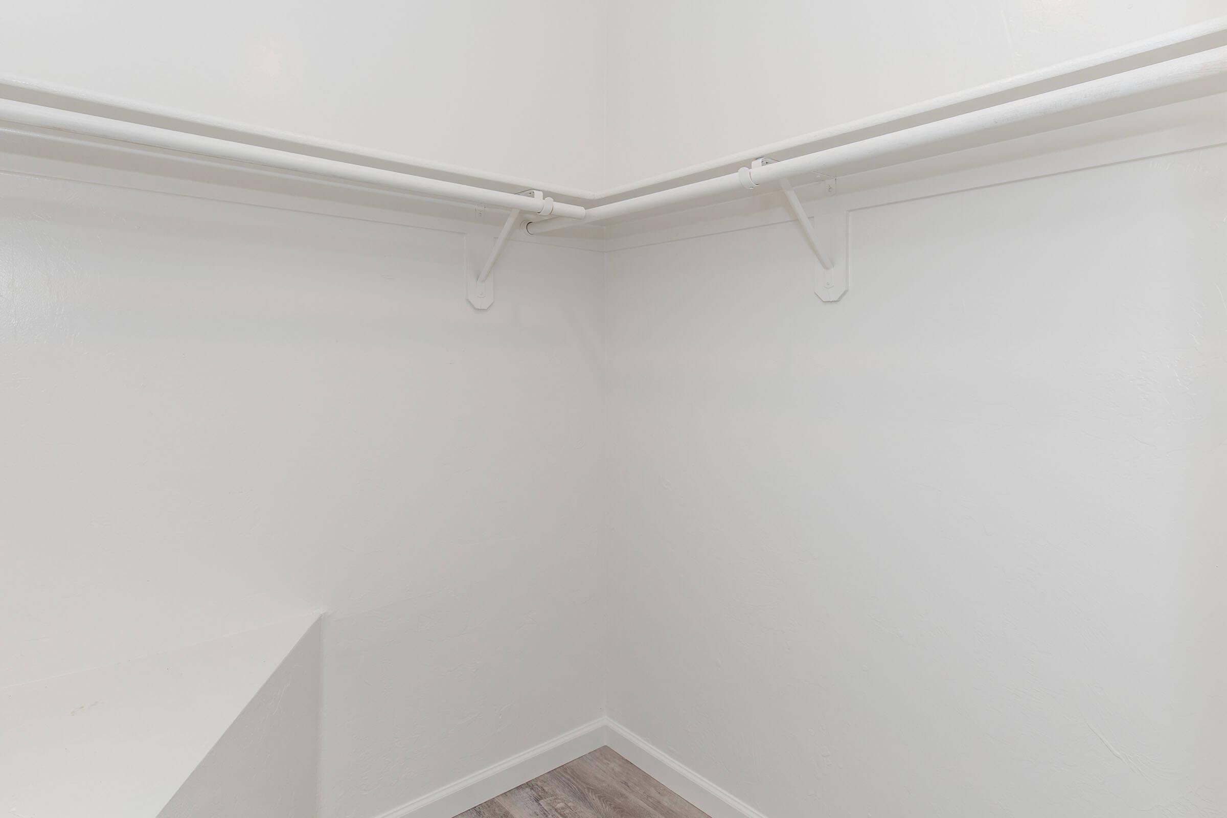 Empty closet with white walls and a light wooden floor. Two white horizontal rods for hanging clothes are installed in the corner. The space appears clean and uncluttered, emphasizing minimalism.