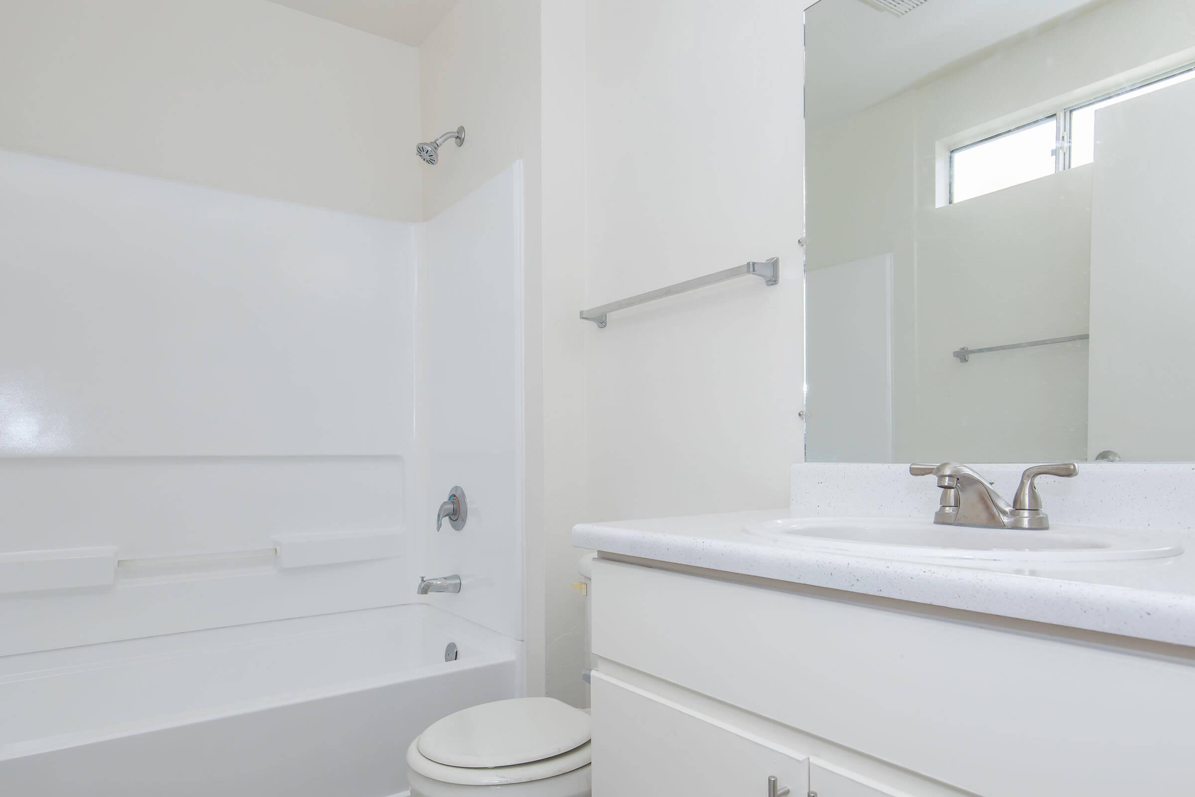 A clean, modern bathroom featuring a white bathtub with a shower, a white sink with a chrome faucet, and a large mirror above the sink. Light-colored walls and a window provide natural light, while a towel bar is fixed on the wall. The overall design is simple and contemporary.