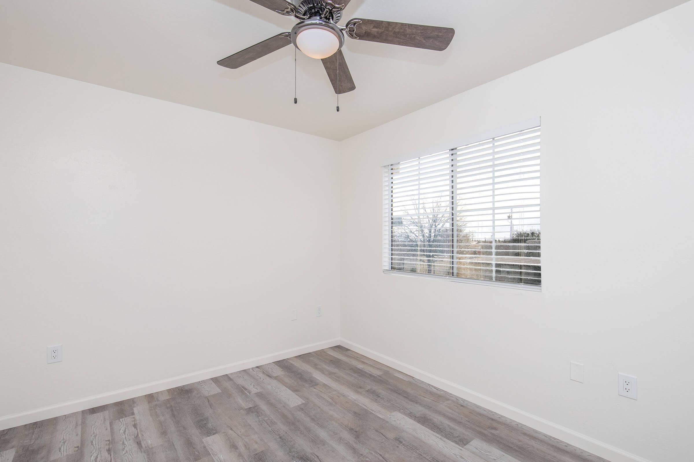 A bright, empty room with light-colored walls and a ceiling fan. There is a window with blinds allowing natural light to enter, and the floor is covered with wooden planks. The room appears clean and ready for furnishing.