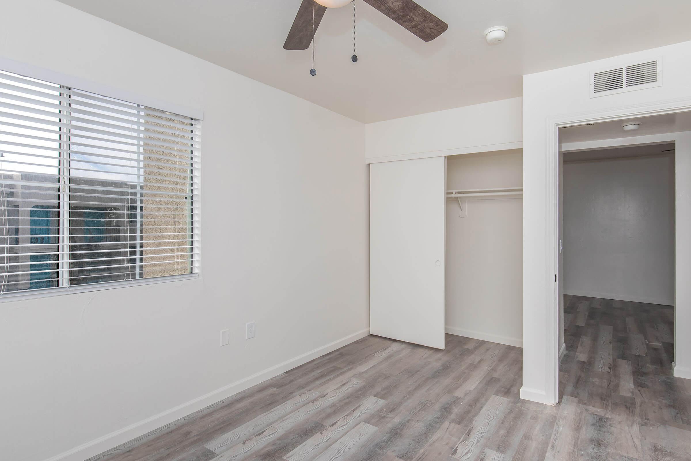A bright, empty bedroom featuring a light-colored wall and wooden flooring. There's a ceiling fan with wooden blades, a closet with sliding doors, and a window with horizontal blinds allowing natural light to enter. The room has a clean and modern appearance.