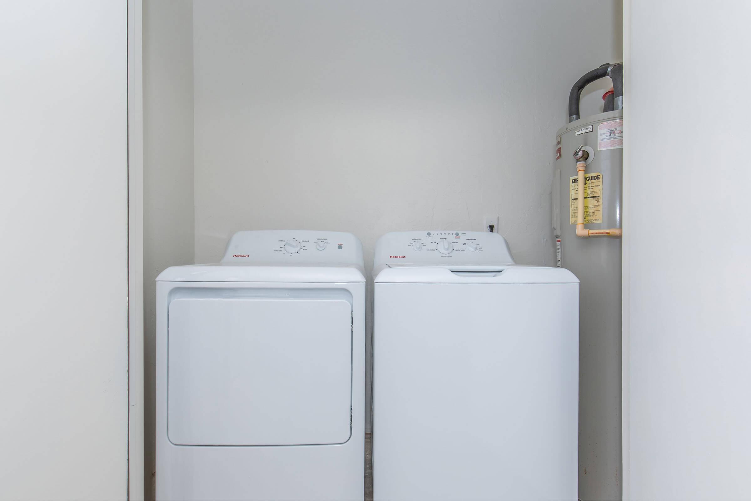 A compact laundry room featuring a white washing machine and dryer side by side, with a water heater mounted on the wall beside them. The room has a light-colored wall and a clean, minimalist design.