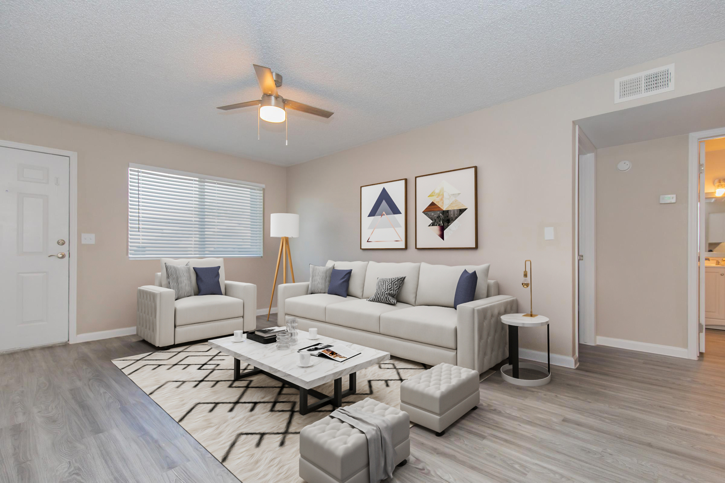 A modern living room featuring a light-colored sofa with blue and white throw pillows, two chairs, a coffee table with magazines, and a geometric area rug. There is a ceiling fan and wall art above the sofa. The room has a neutral color palette and a bright window letting in natural light.
