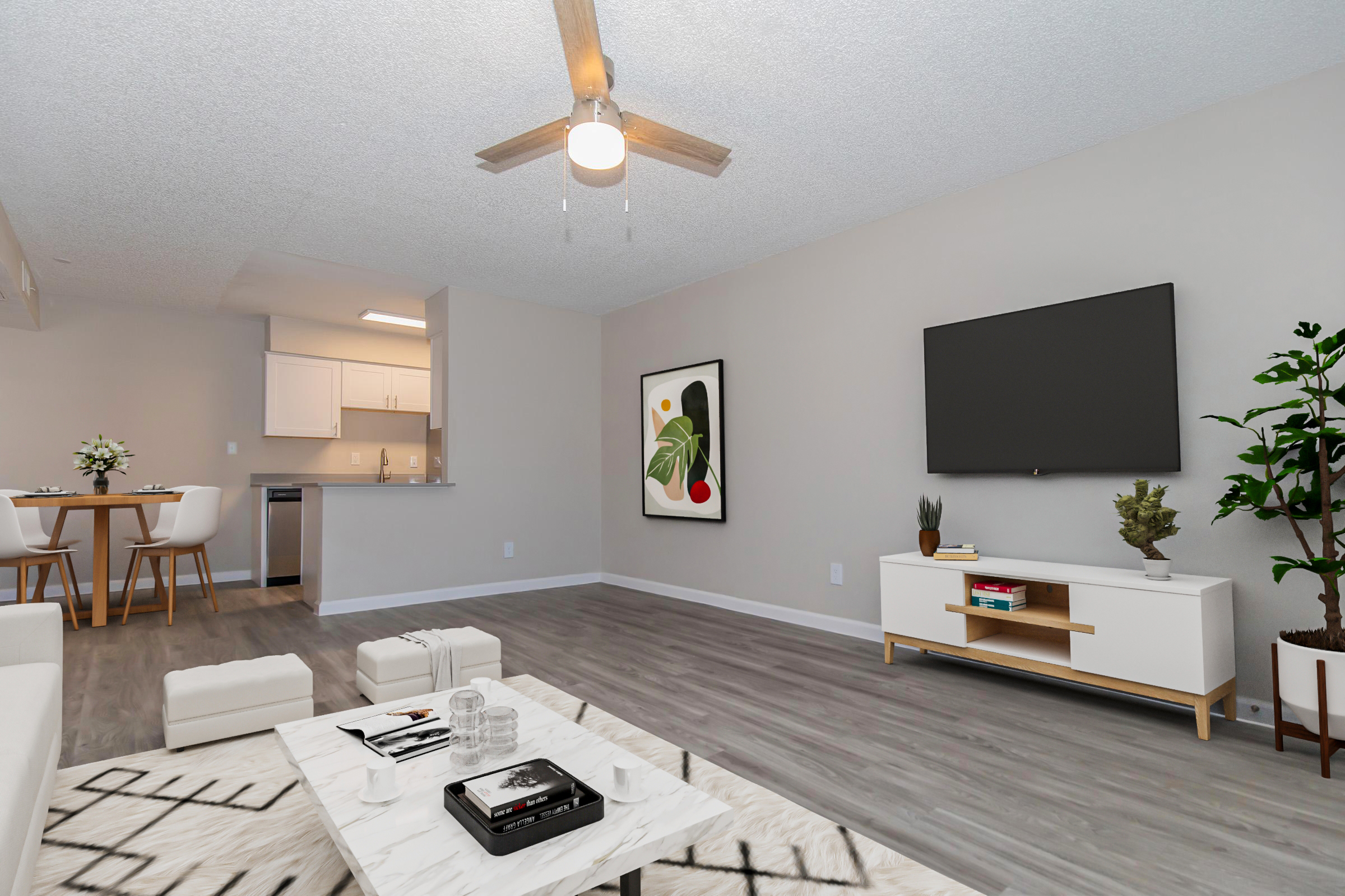A modern living room featuring a gray ceiling, a wooden fan, light-colored walls, and a large TV mounted on the wall. A coffee table with books and decor items sits on a wooden floor, while a potted plant adds greenery. In the background, there's a dining area with a round table and chairs.
